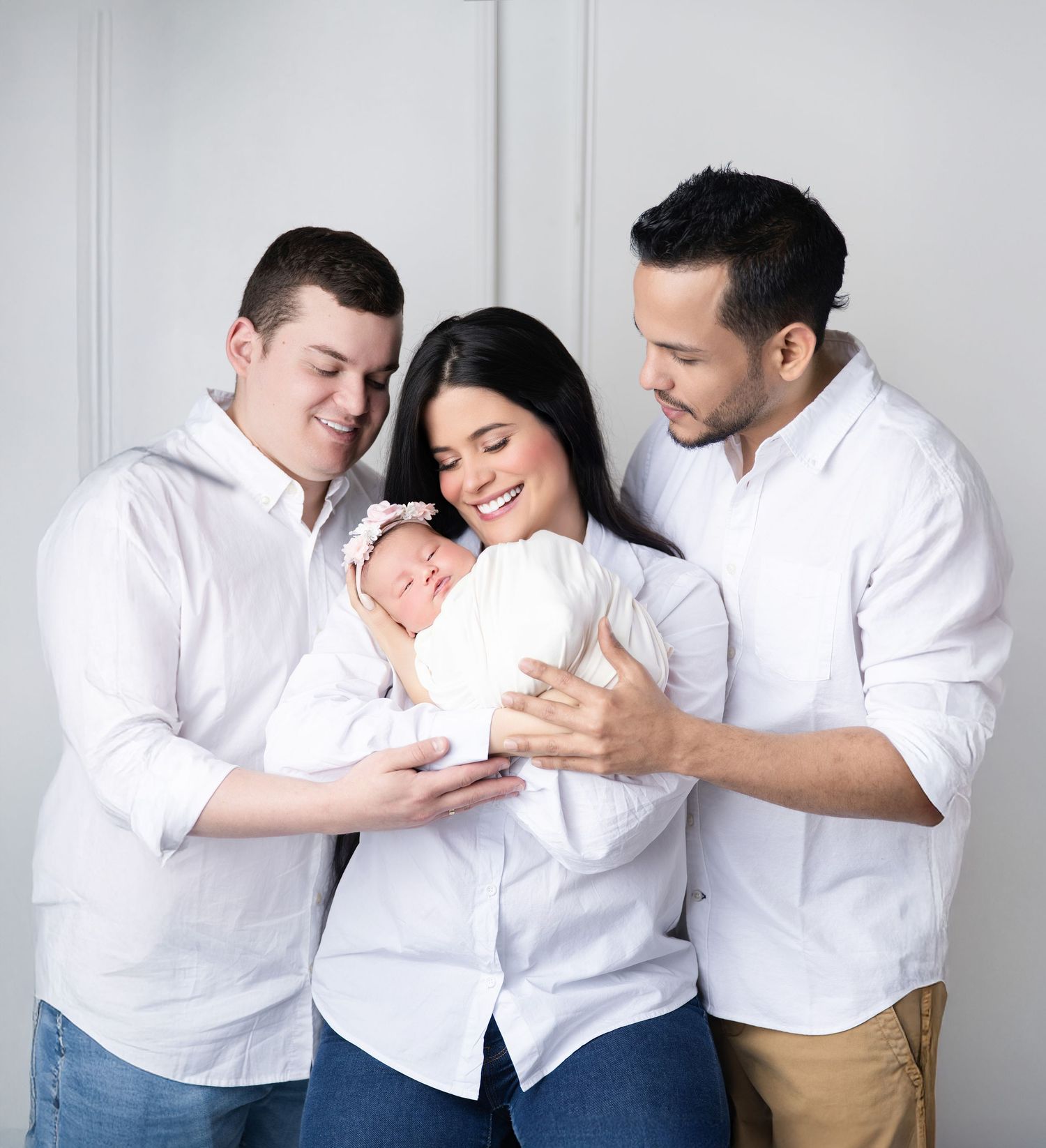 Family of three in white shirts smiling at newborn baby girl in a bright studio portrait.