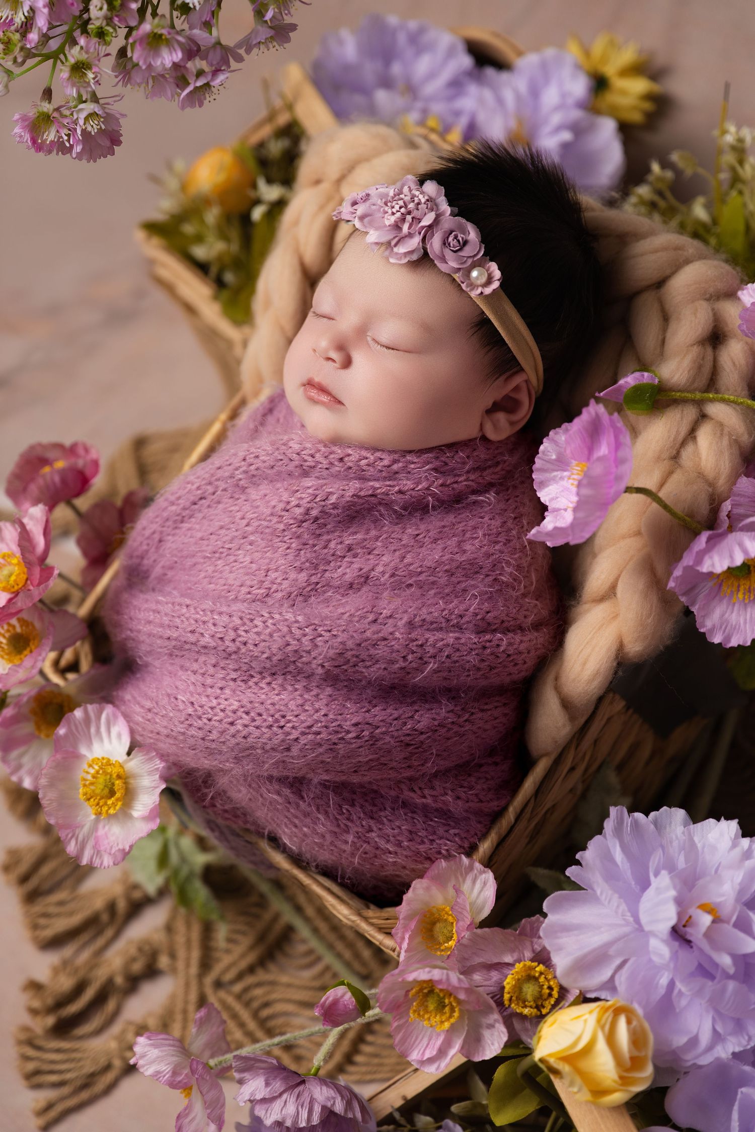 Sleeping newborn baby girl wrapped in purple knit swaddle, wearing floral headband, nestled in basket with blooming flowers.