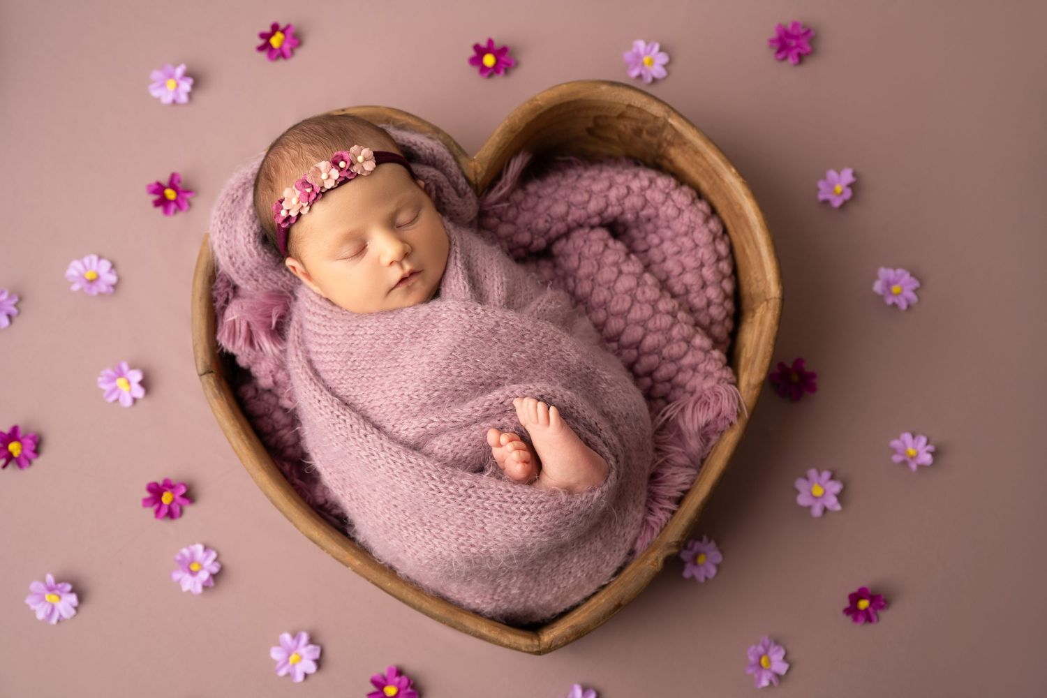 Newborn baby swaddled in pink wrap sleeping in heart-shaped wooden bowl surrounded by purple flowers.