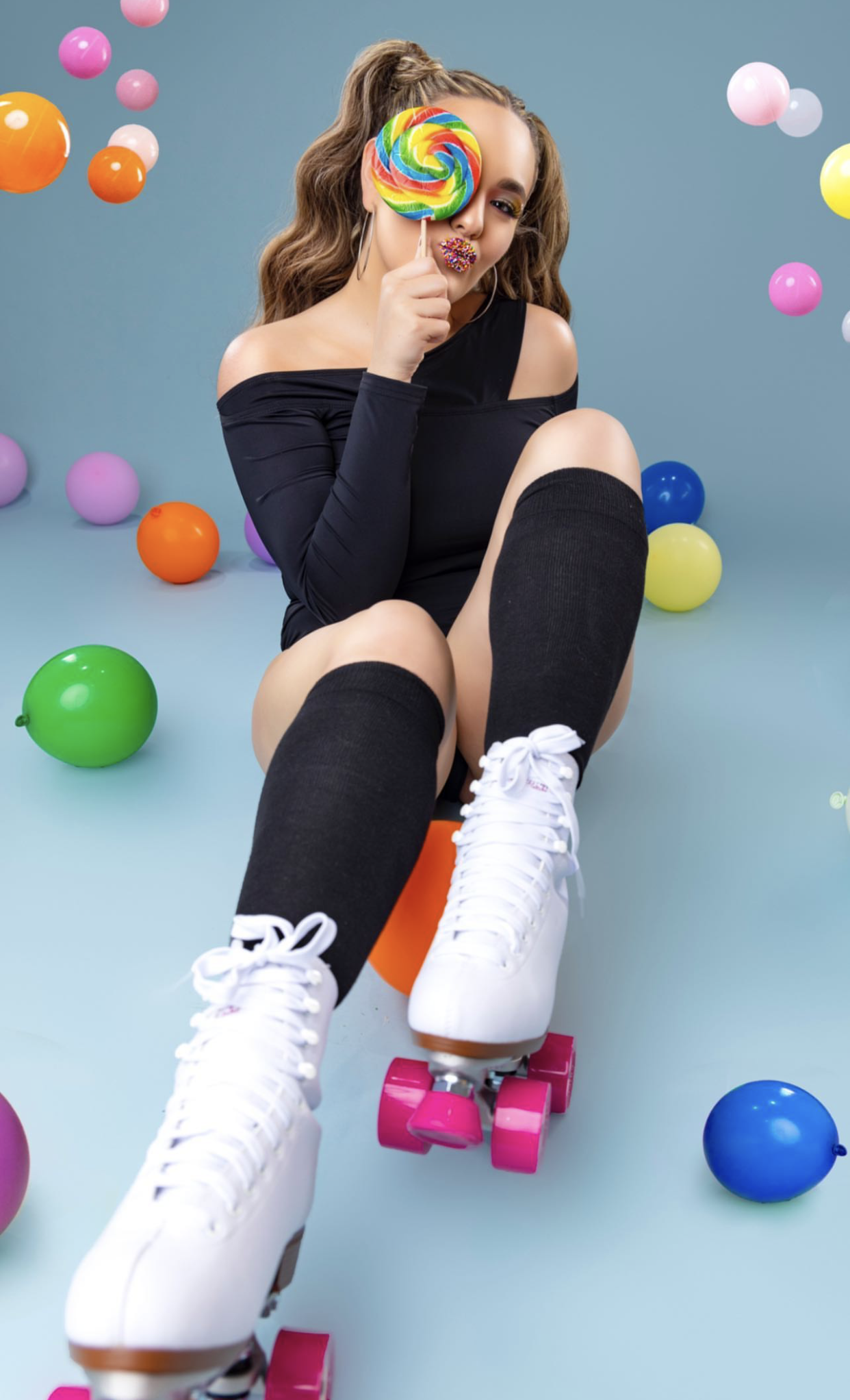 Young woman in black bodysuit and white roller skates holding a colorful lollipop surrounded by vibrant balloons.