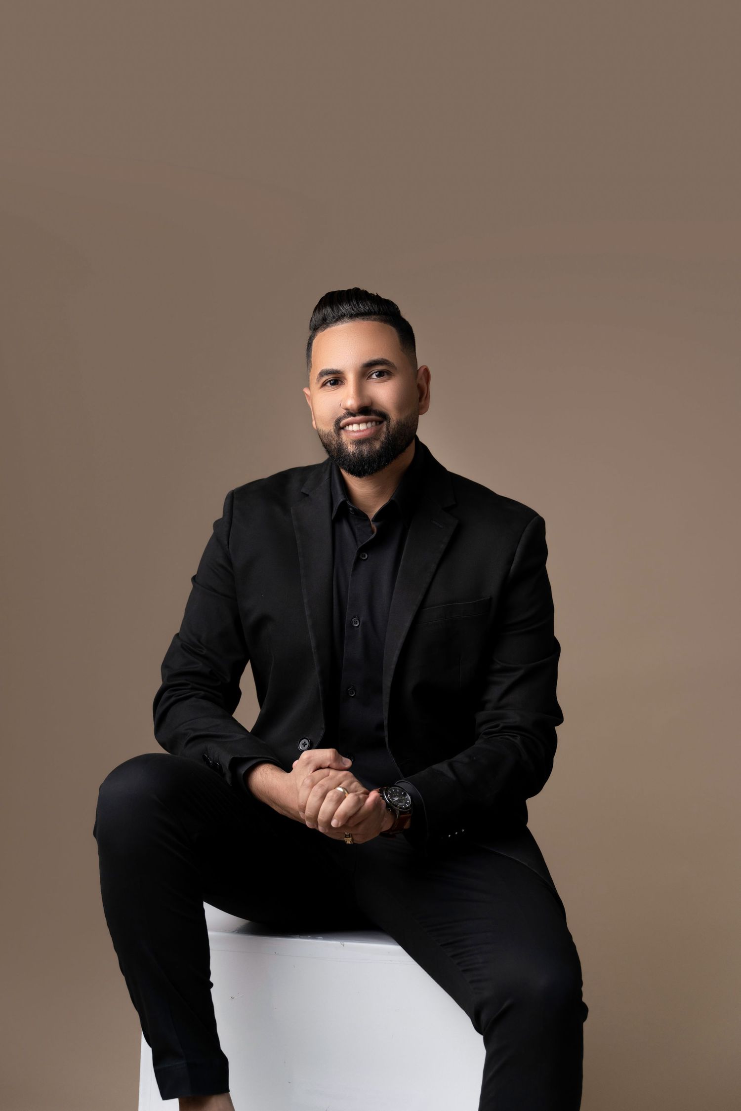 A confident man in a black suit sits on a white cube against a tan background, smiling with hands clasped.