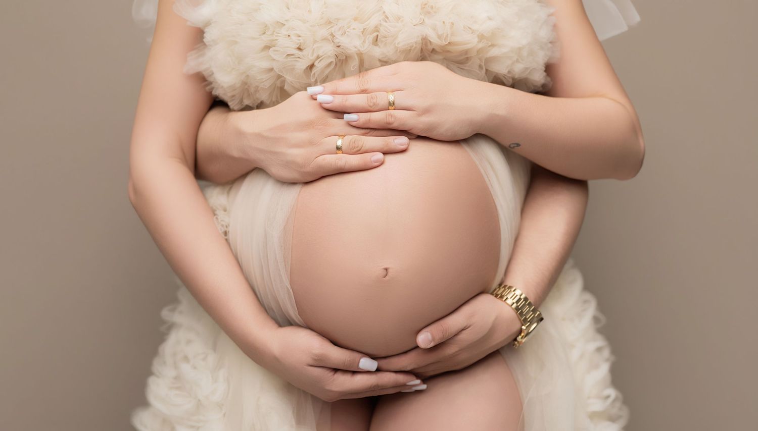 Pregnant woman in white tulle dress cradling bare baby bump with both hands, wearing gold bracelet and ring.