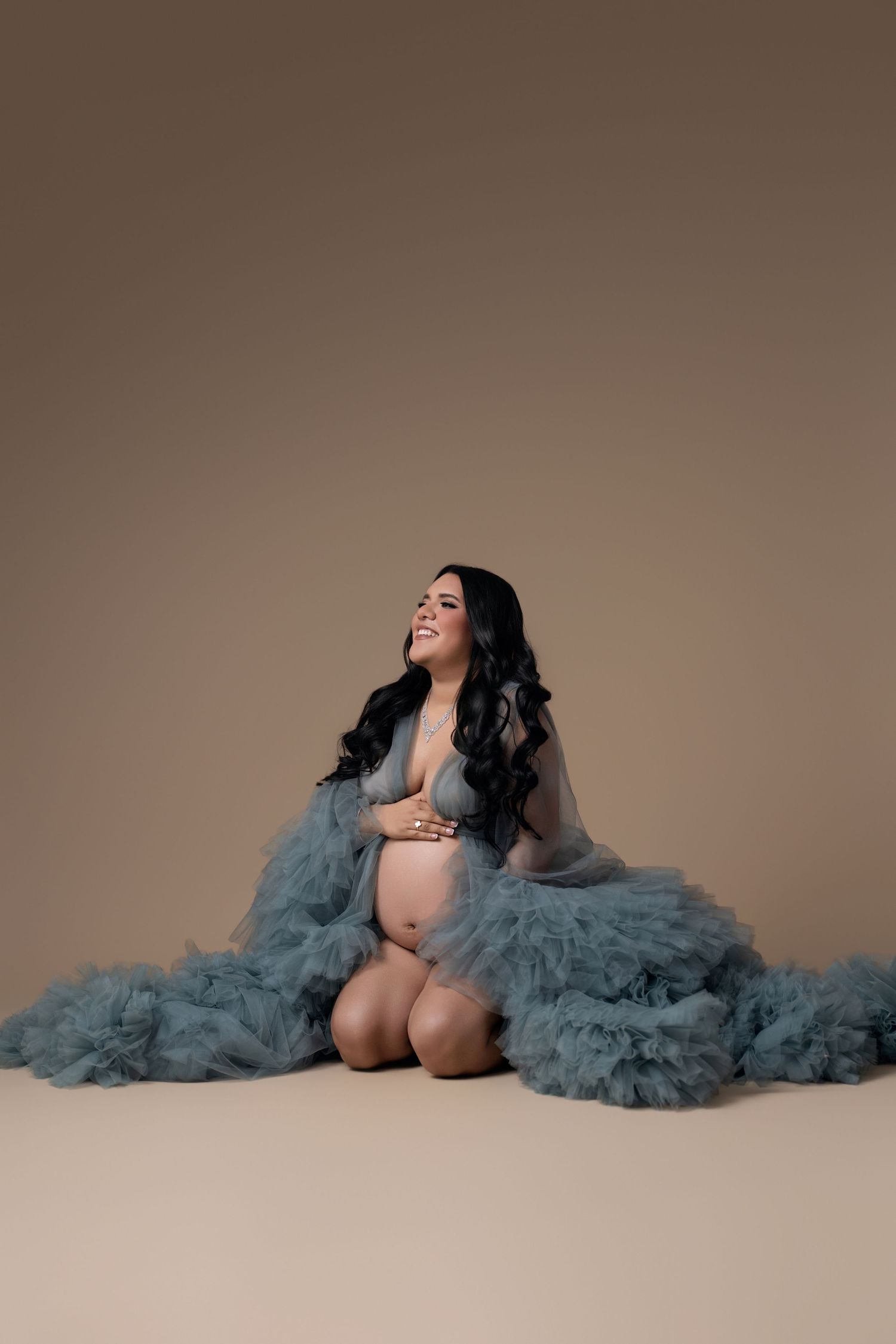 Pregnant woman in blue tulle robe sitting gracefully on studio floor, smiling, with long dark hair against beige background.