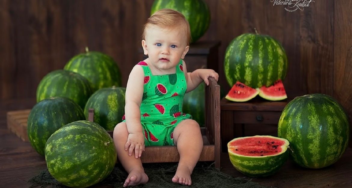 A small child in a watermelon-print romper sits surrounded by fresh watermelons against a rustic wooden backdrop.