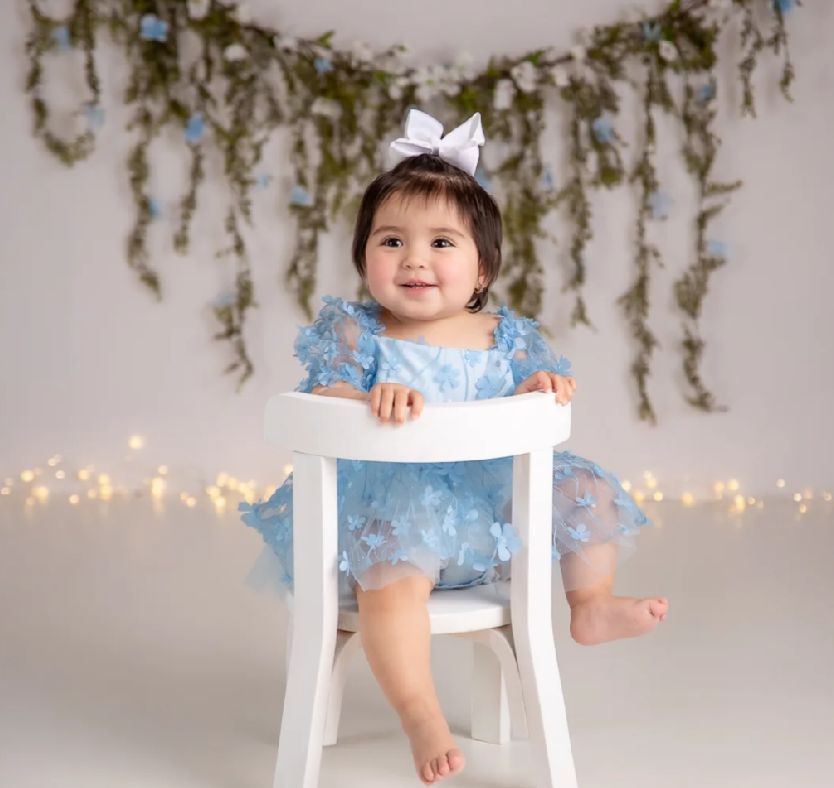 Baby in blue tulle dress and crown sitting on white chair with twinkle lights and greenery backdrop.