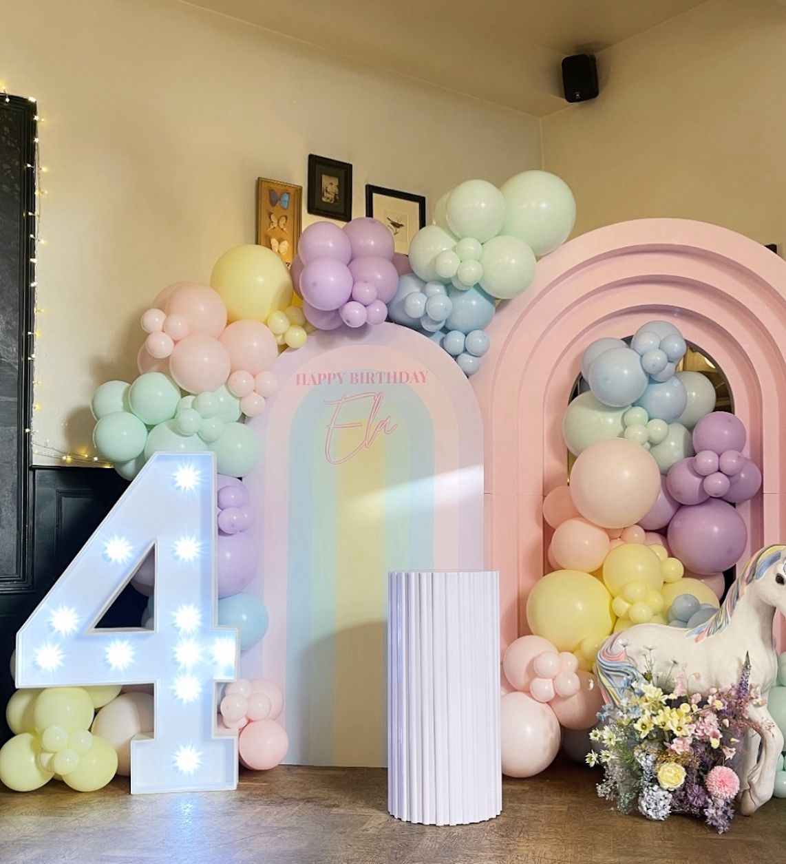 Pastel balloon arch installation with light-up number 4 and columns in soft rainbow colors for birthday party decor.