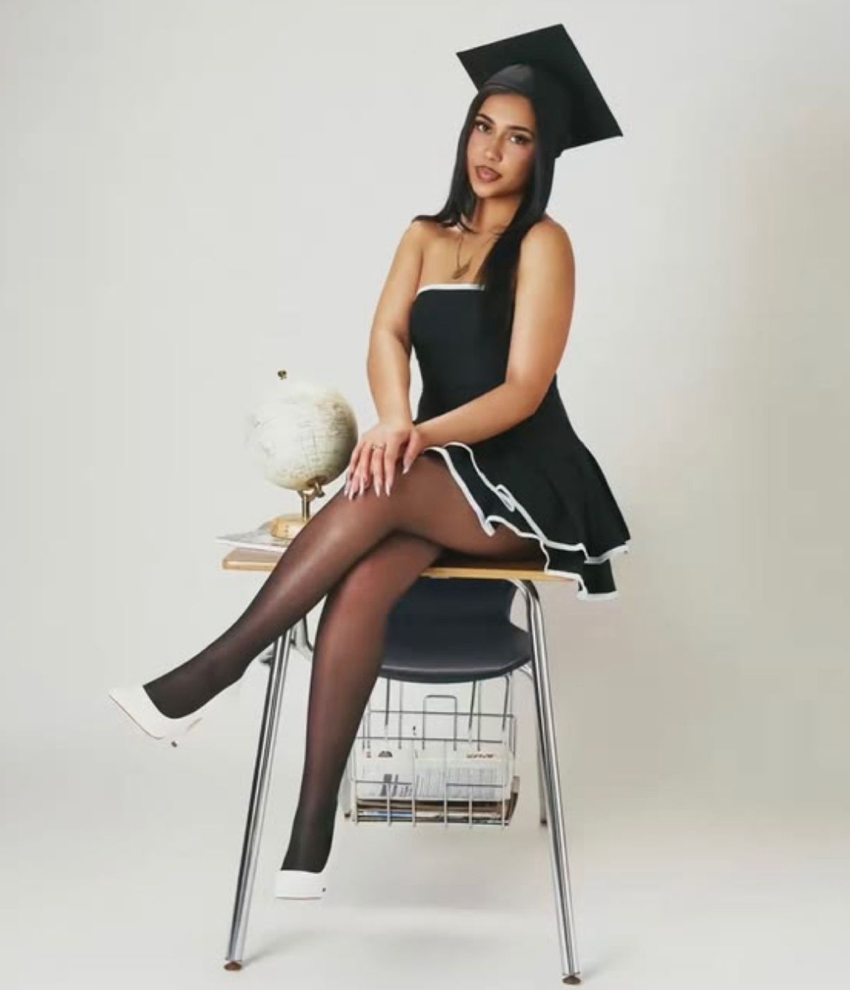 Graduate in black dress and cap poses on modern chair against white backdrop for graduation photos.