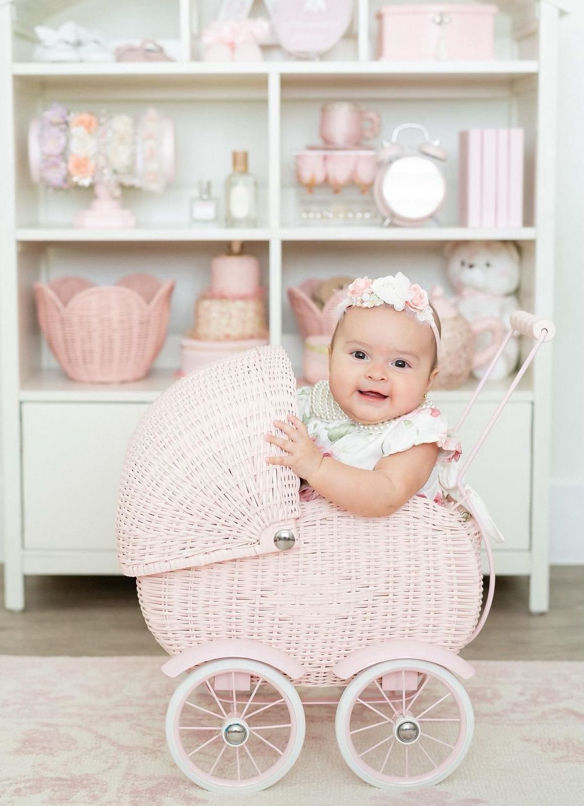 Pink vintage-style wicker baby carriage with decorative shelving display in pastel nursery setting.