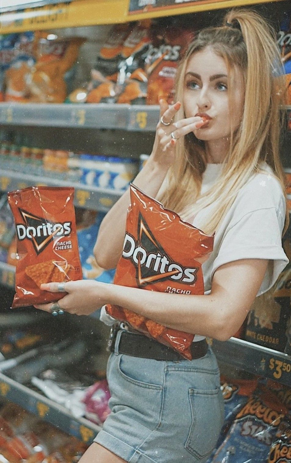 Young woman in white tee and denim shorts holding Doritos nacho cheese chips bags in a snack aisle.