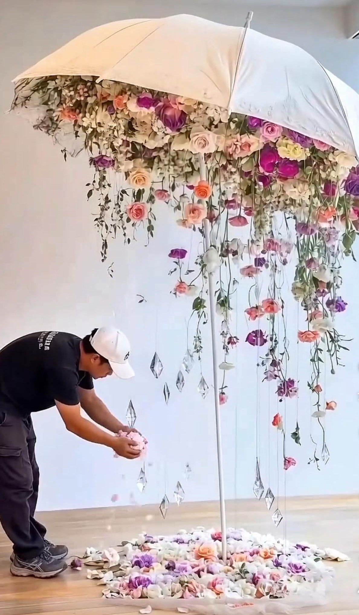 Artist arranges colorful flowers cascading from a large white umbrella installation with petals scattered on the floor.