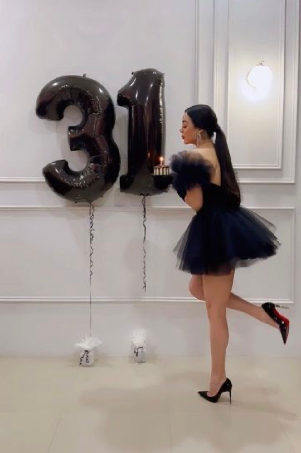 Woman in black tutu dress holding birthday cake beside number 31 balloons, wearing red-sole heels.