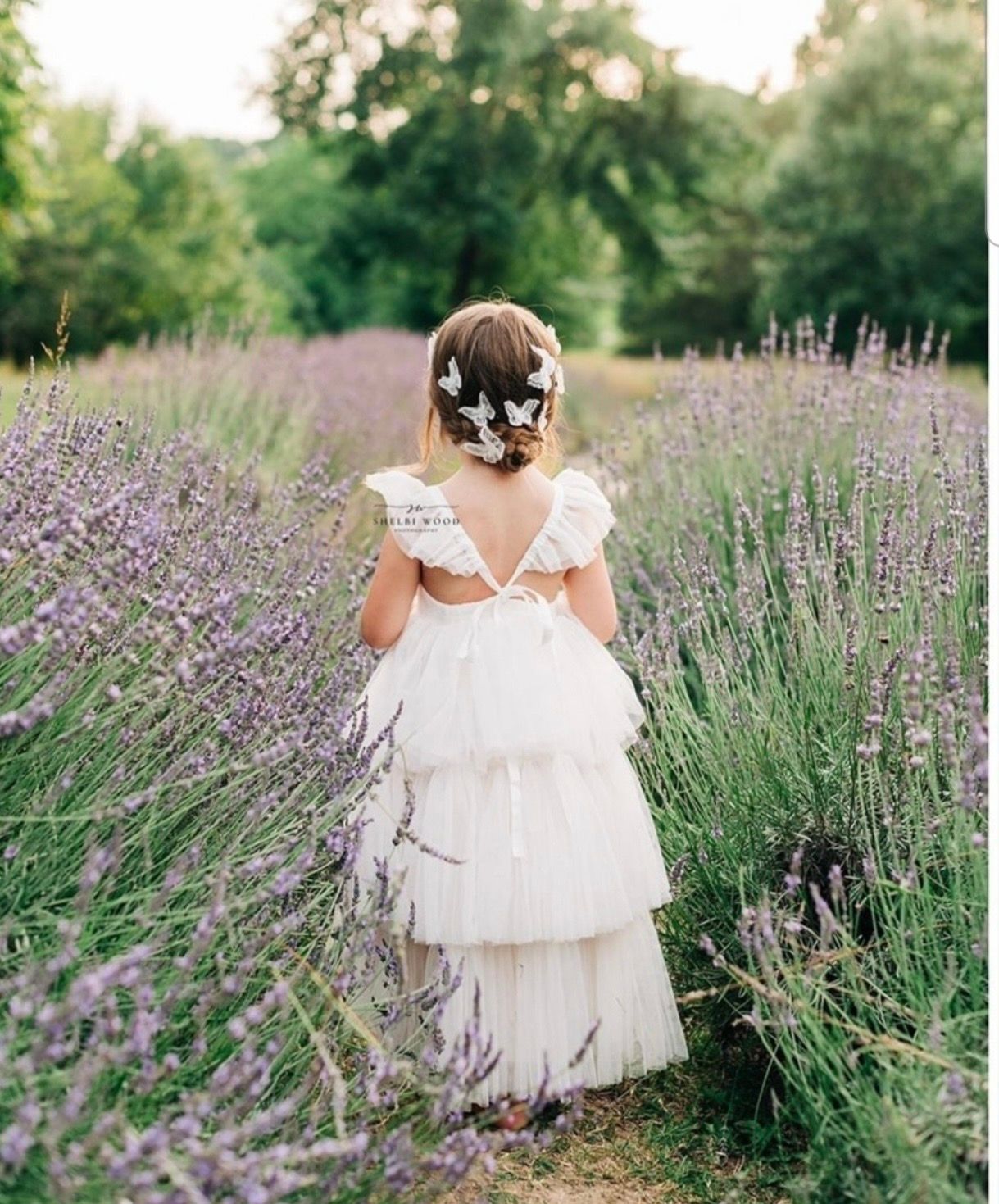 Young flower girl in white tiered dress walks through lavender field with butterfly hair accessories.