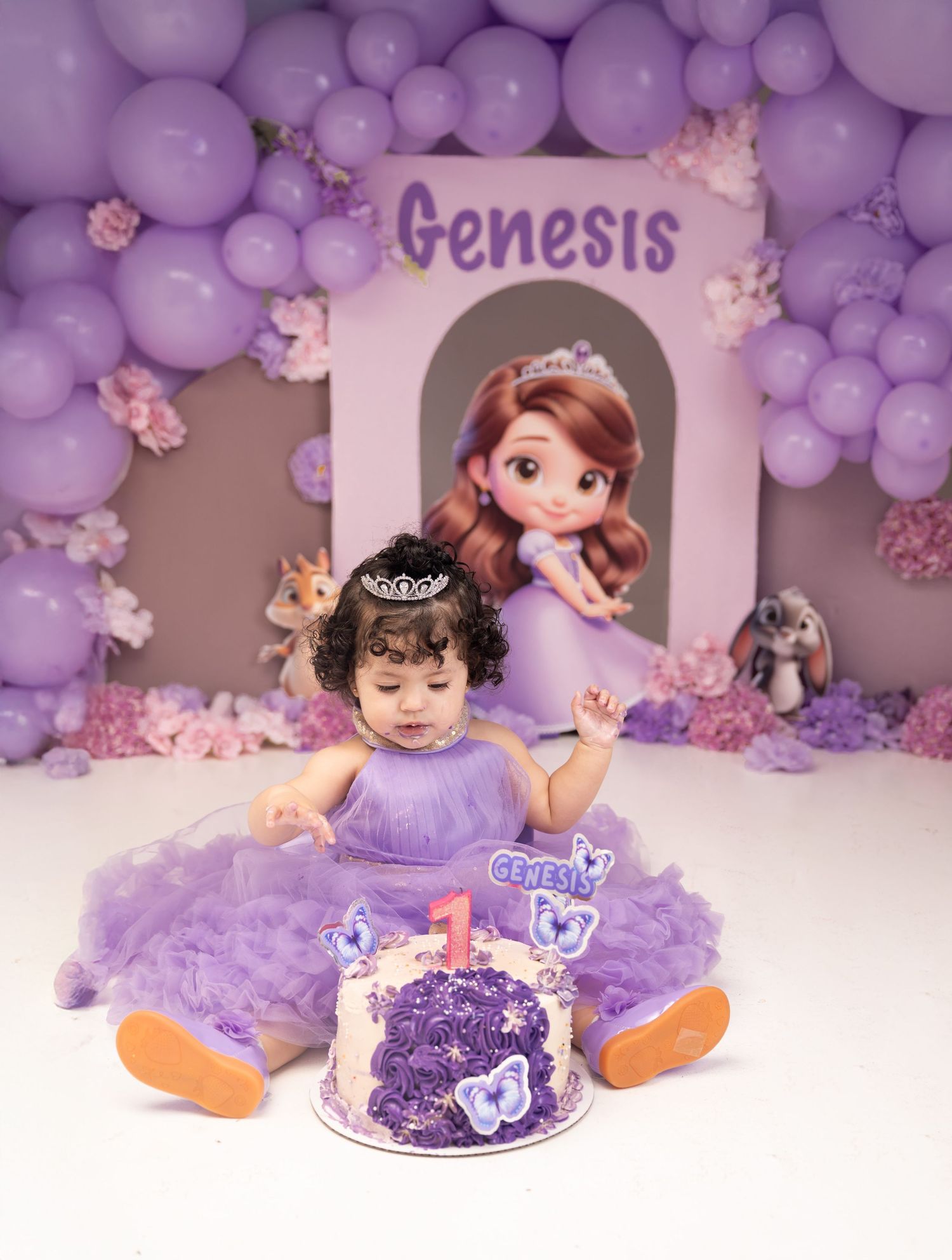 Baby girl in purple tutu dress celebrates first birthday with Sofia the Princess themed cake and balloon backdrop.