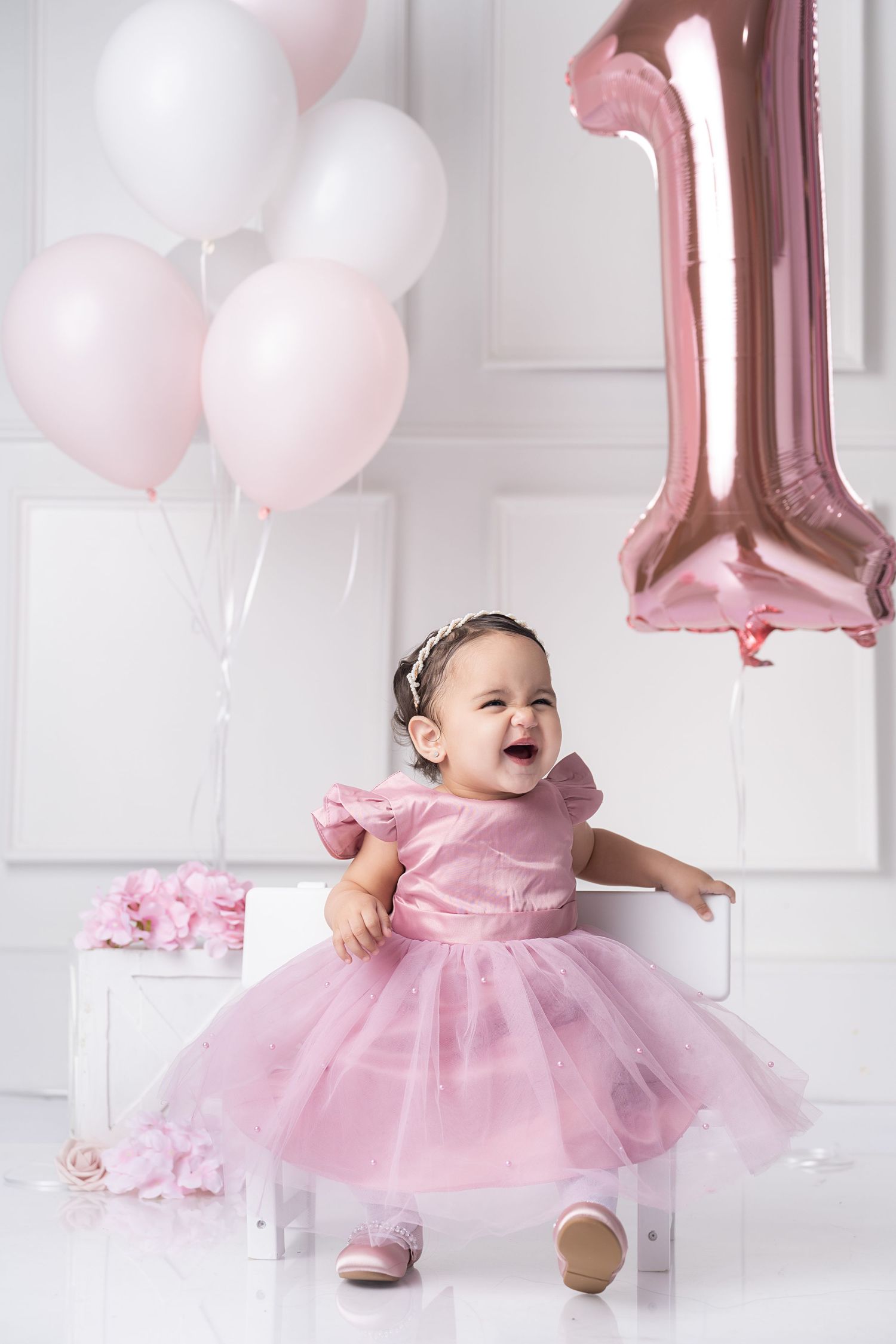 A joyful baby girl in a pink tutu dress celebrates her 1st birthday with balloons in a white studio setting.