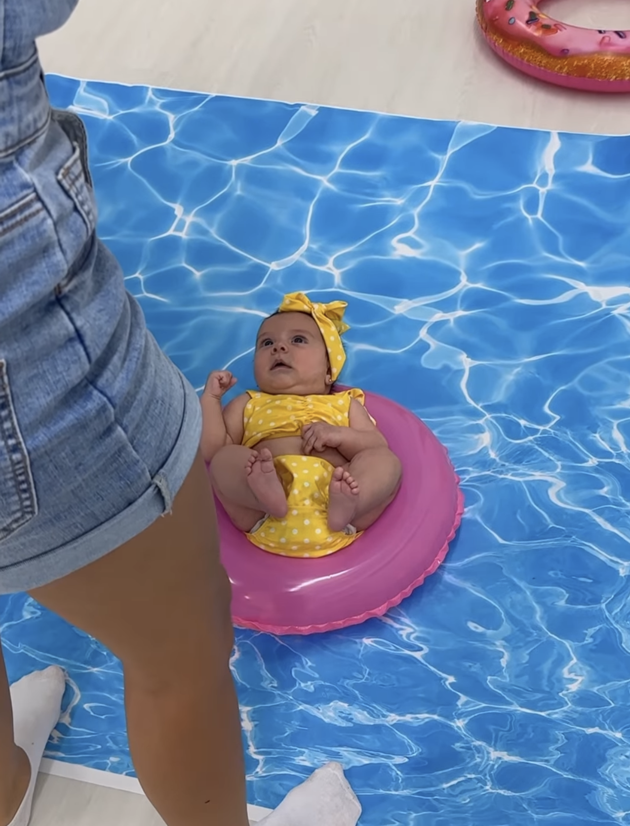 Baby in yellow outfit sitting in pink inflatable ring on pool-themed photo backdrop mat.