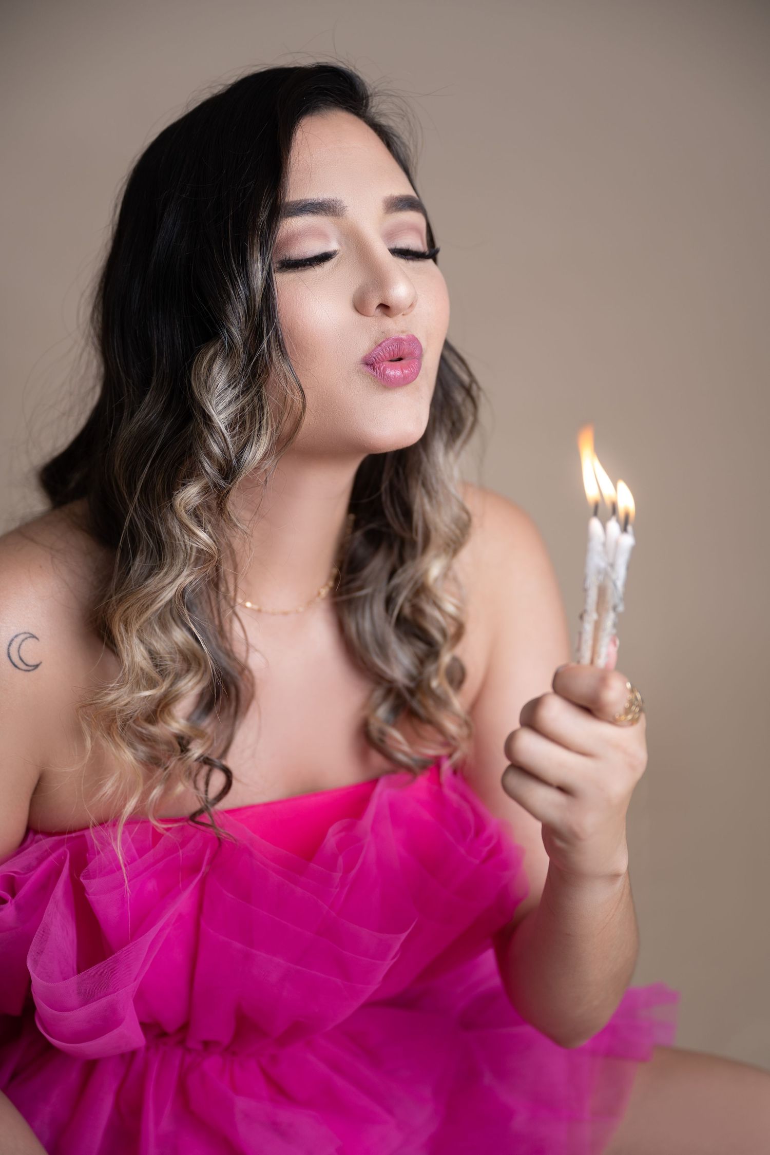 Woman in a hot pink tulle dress with wavy hair closes her eyes and blows out lit birthday candles in a studio portrait.