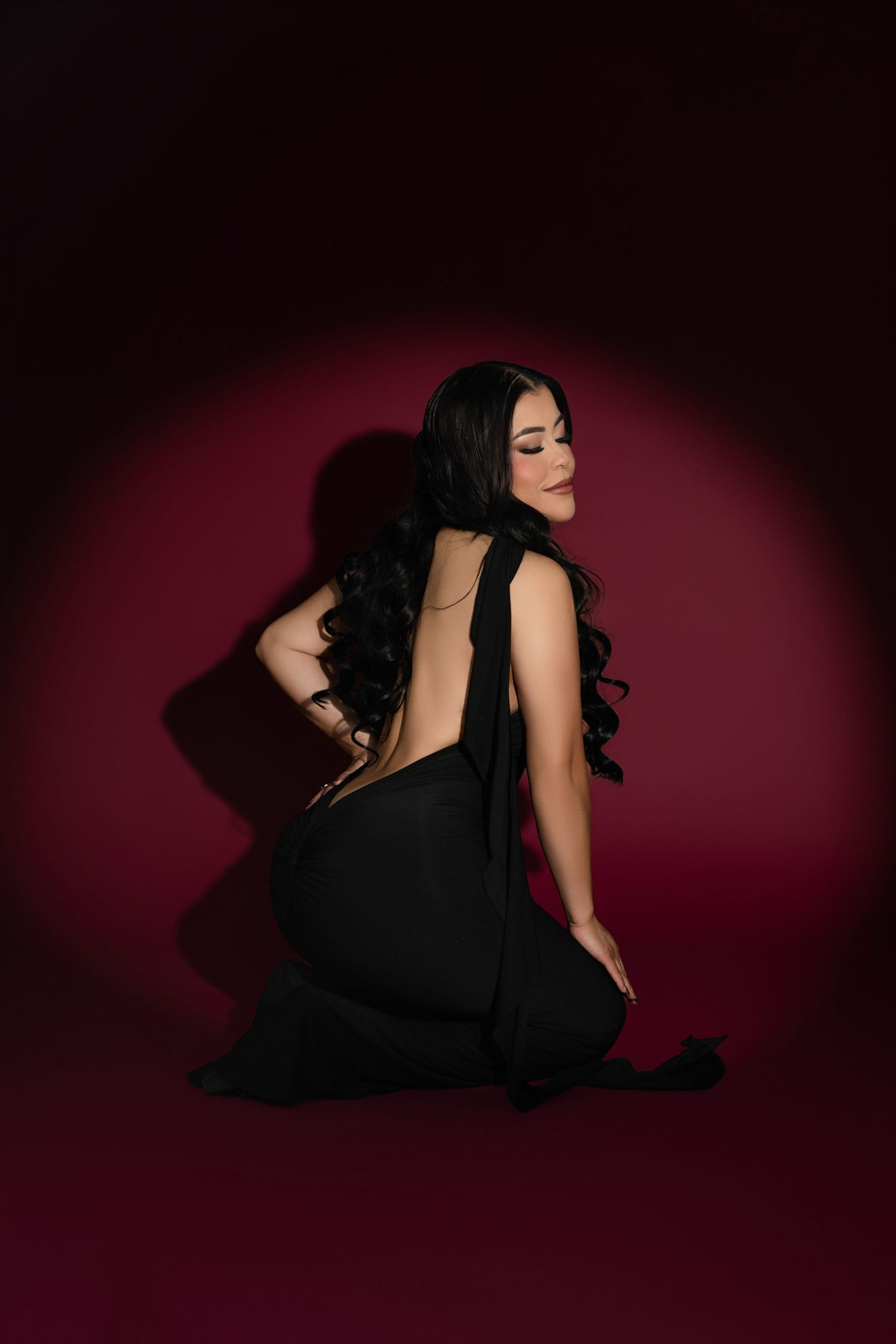 Woman in backless black gown sitting gracefully on floor against deep red backdrop with dramatic lighting.