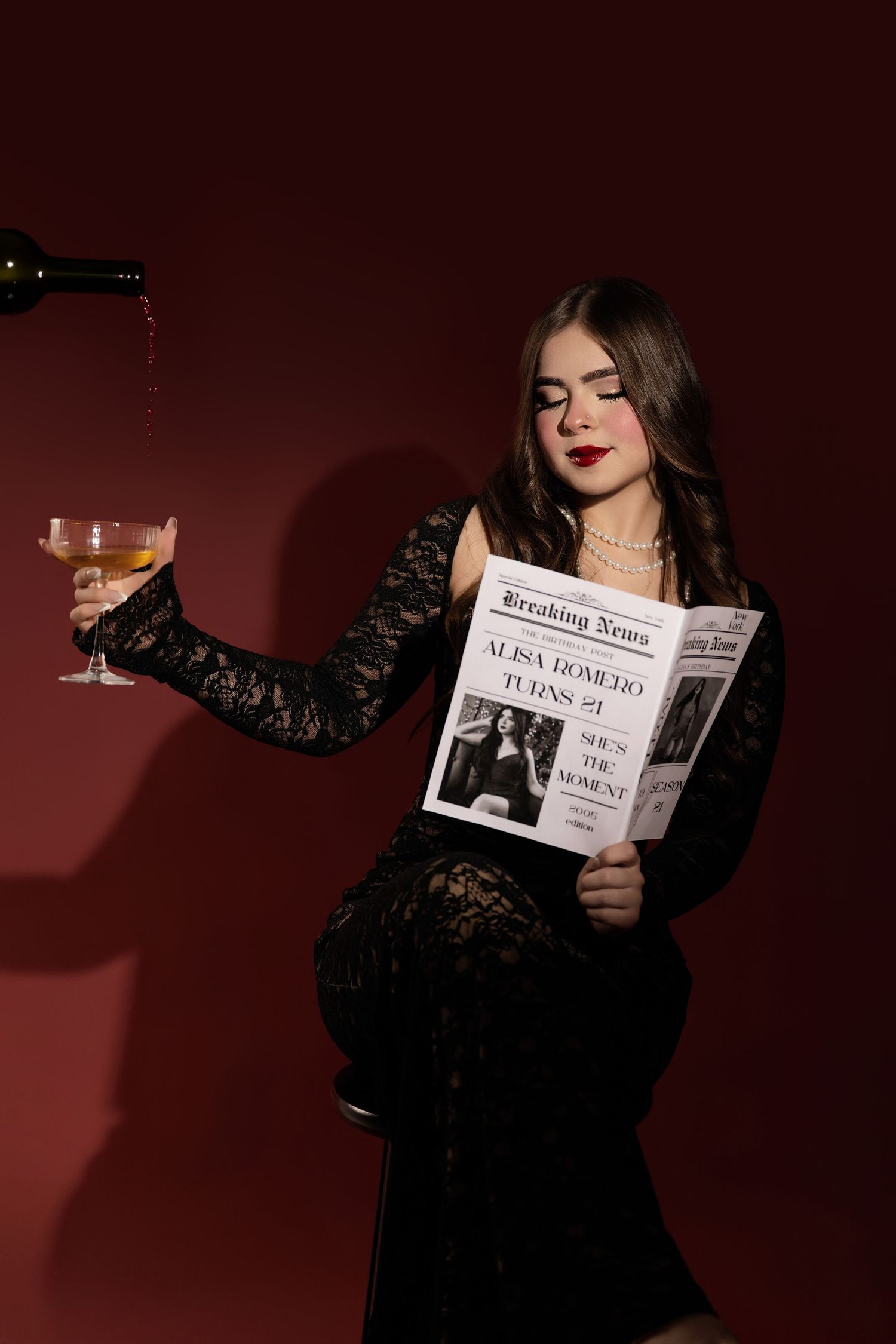 Elegant woman in black lace dress holding wine glass while reading newspaper, wine pouring, dark red background.