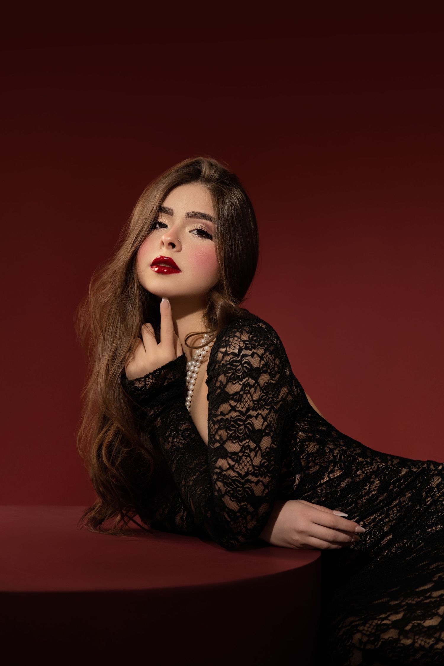 A woman in a black lace dress with pearl necklace poses elegantly against a deep red background with bold red lips.
