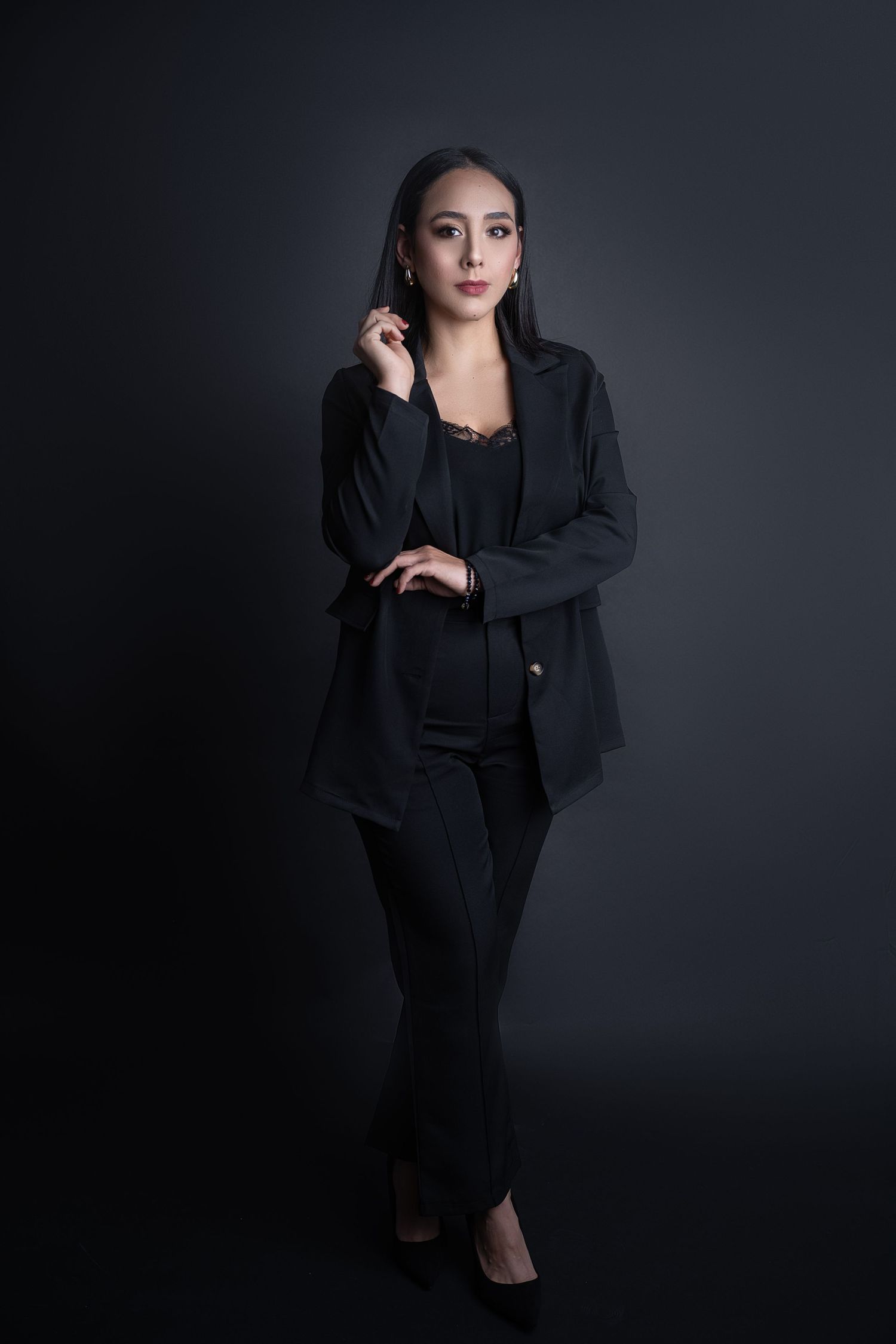 Woman in a black blazer, lace camisole, and tailored trousers poses with her hand near her ear against a dark studio backdrop.