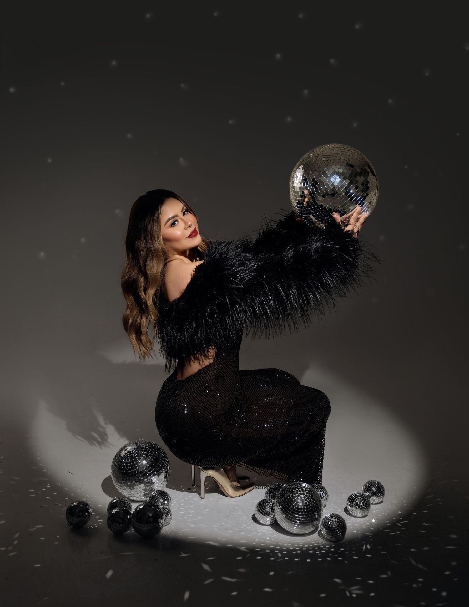 Woman in a black feather jacket and sparkly dress crouches in a spotlight, holding a mirrored disco ball surrounded by smaller disco balls.