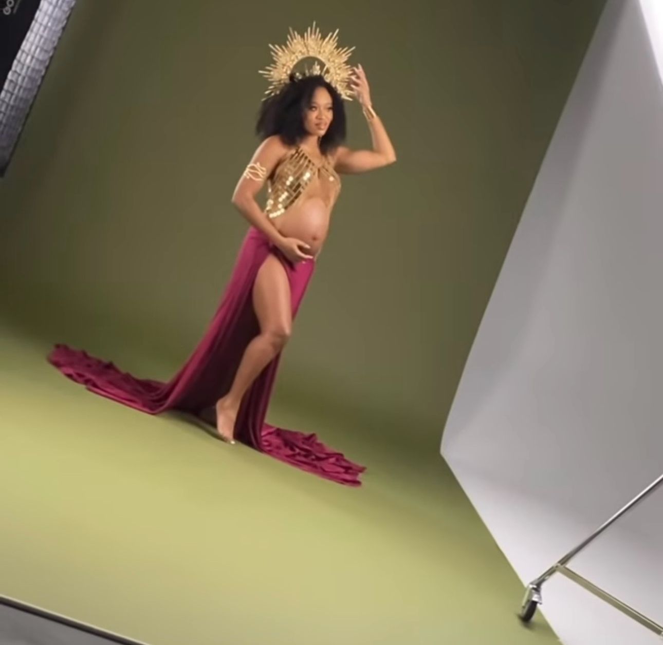 Pregnant woman in gold crown and magenta gown posing in a professional photo studio setting.