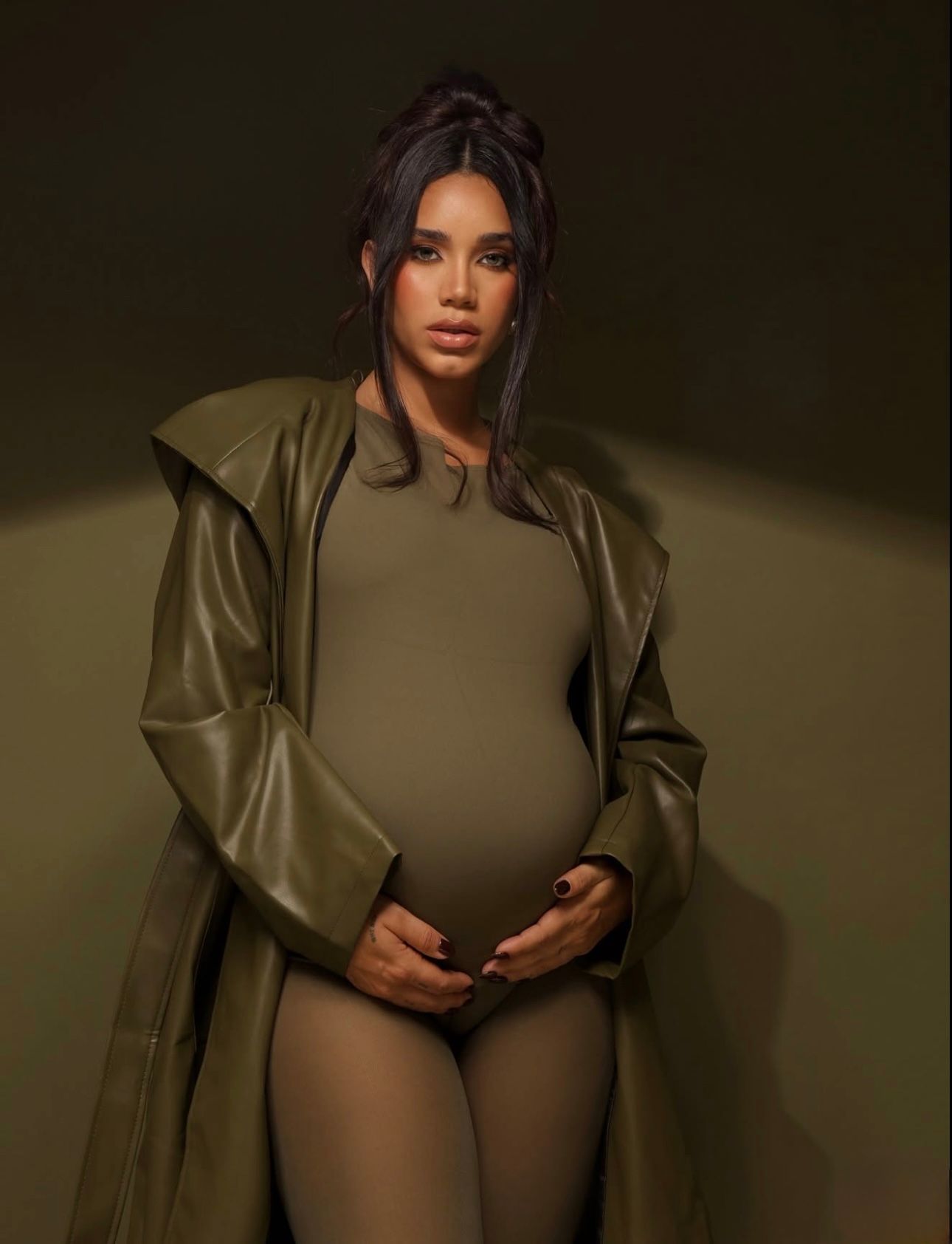 Pregnant woman in olive green bodysuit and leather trench coat cradling baby bump in a moody studio setting.
