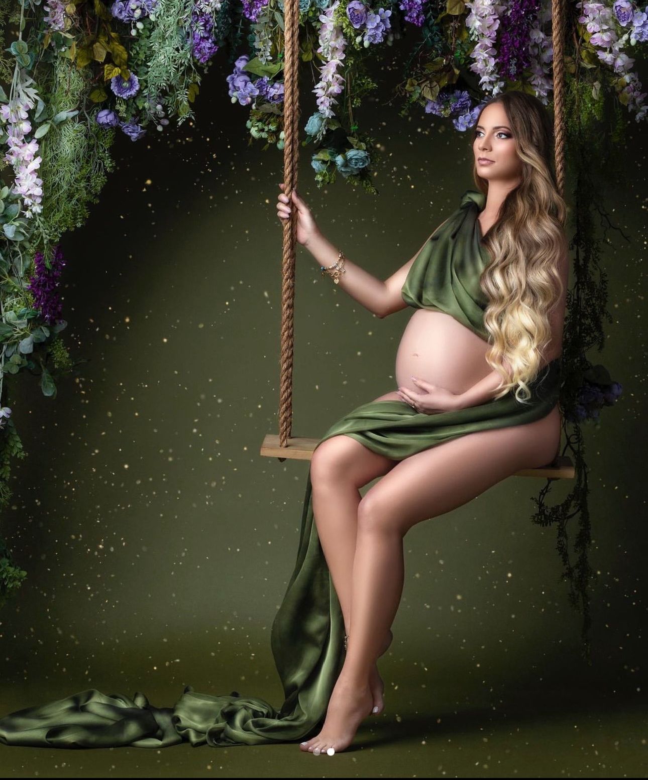 Pregnant woman in green silk wrap sitting on a rope swing surrounded by lush floral backdrop for maternity shoot.
