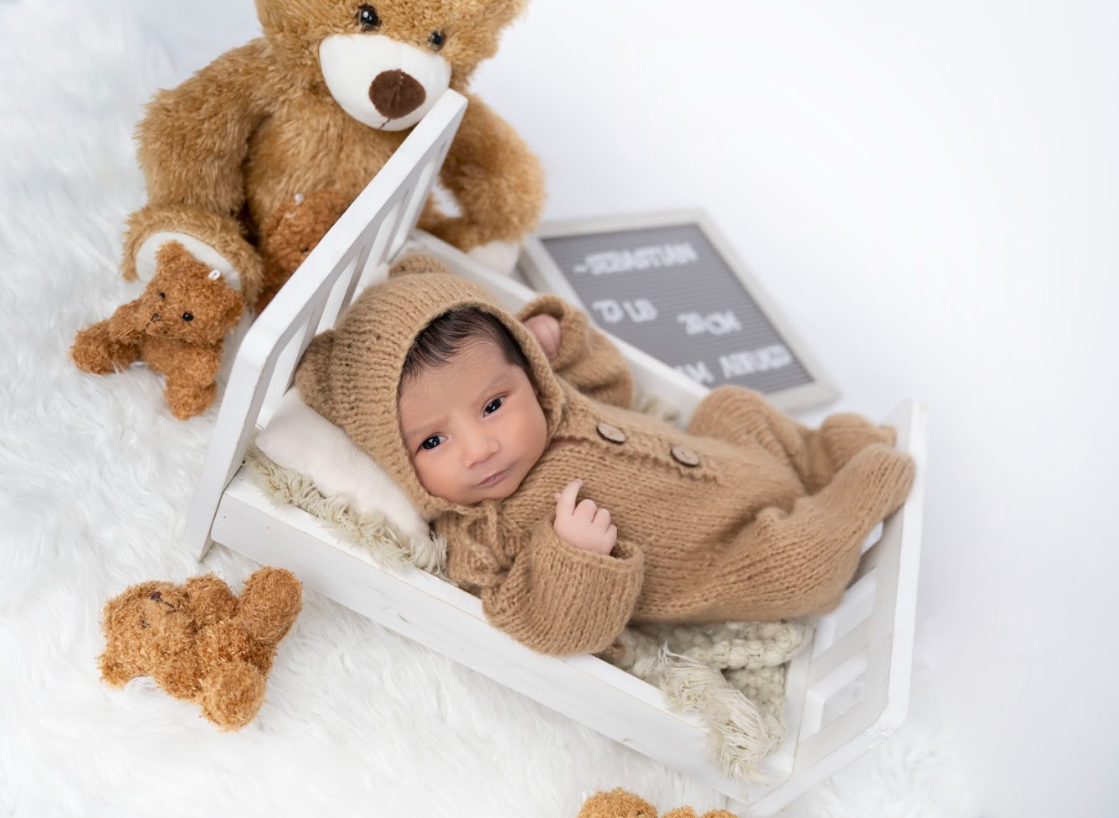 A baby in a cozy brown knit sweater lies on a soft blanket next to plush teddy bears.