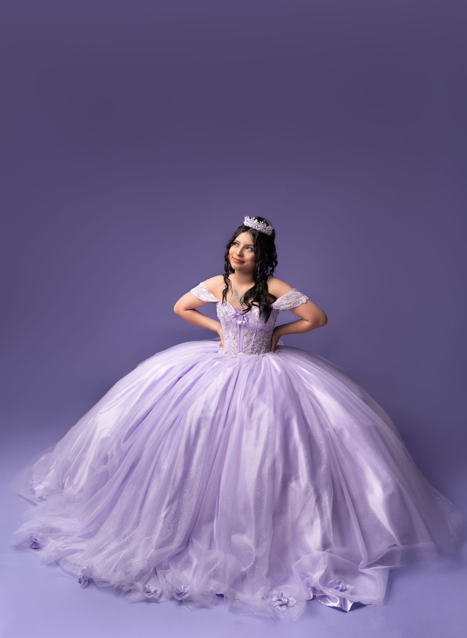Young woman in lavender ball gown with tiara poses gracefully for quinceañera portrait against purple background.