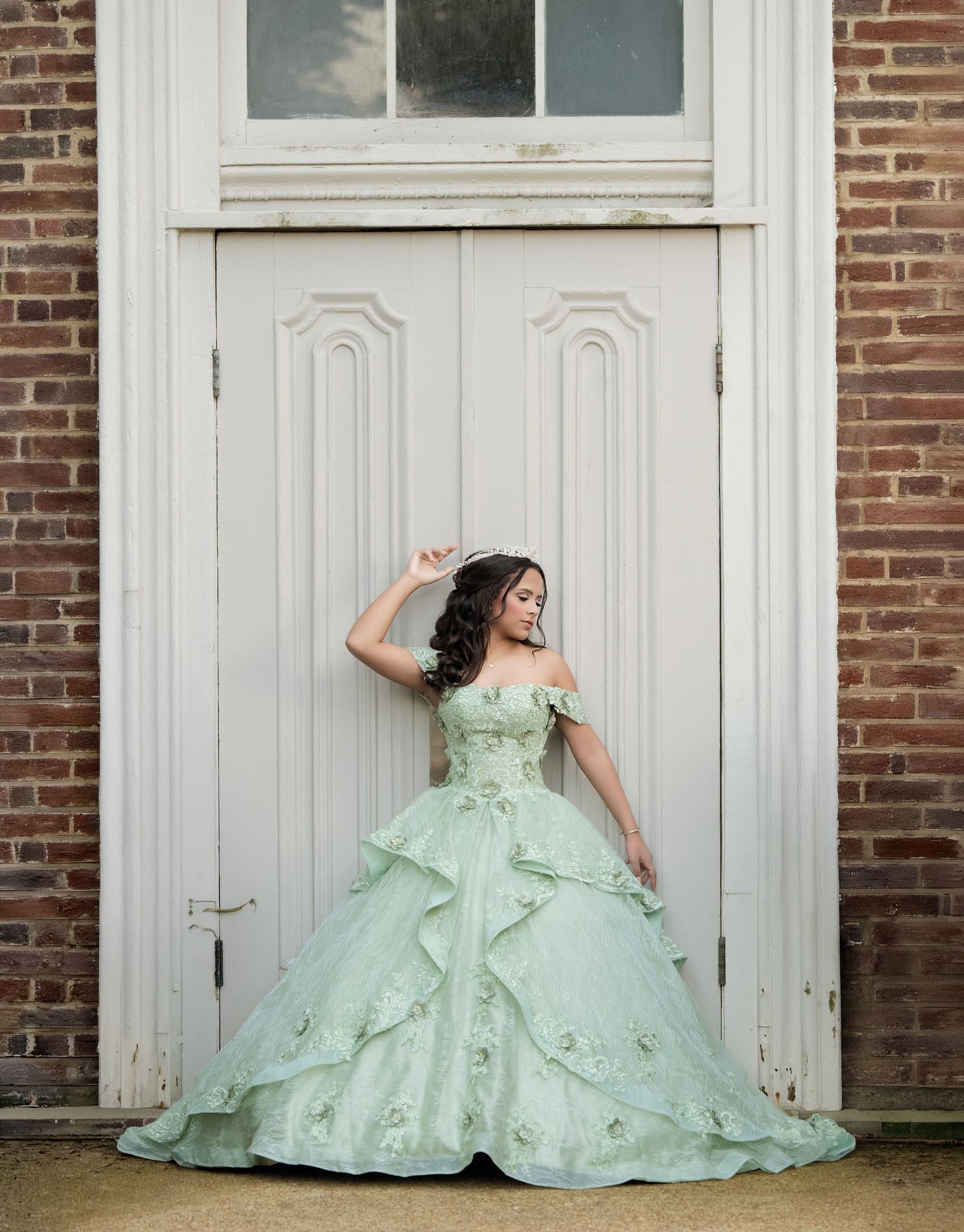Young woman in mint green off-shoulder ball gown with floral details poses elegantly before a white door.