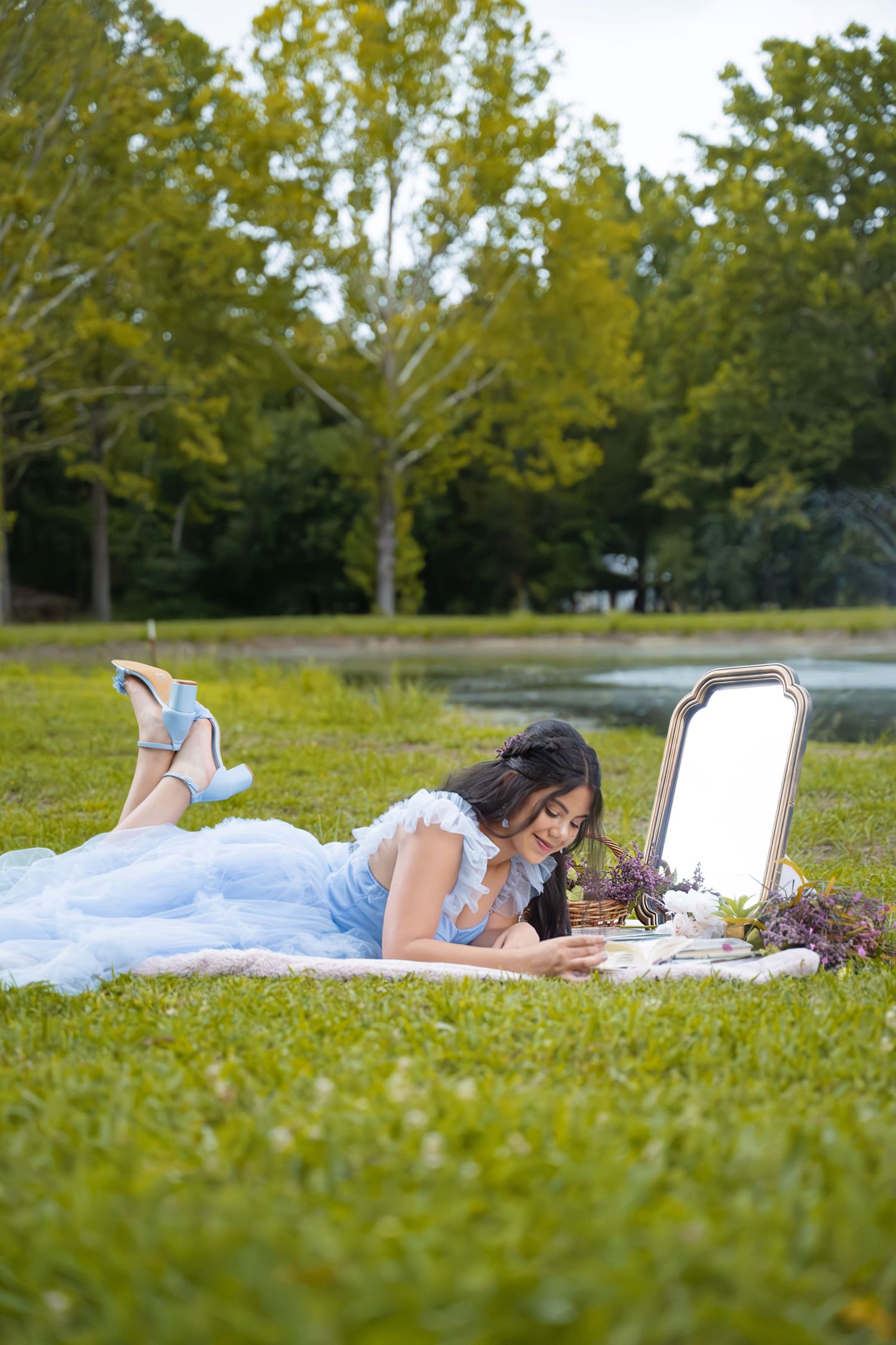 Young woman in blue gown lying on grass in park, reading beside vintage mirror and flower basket.