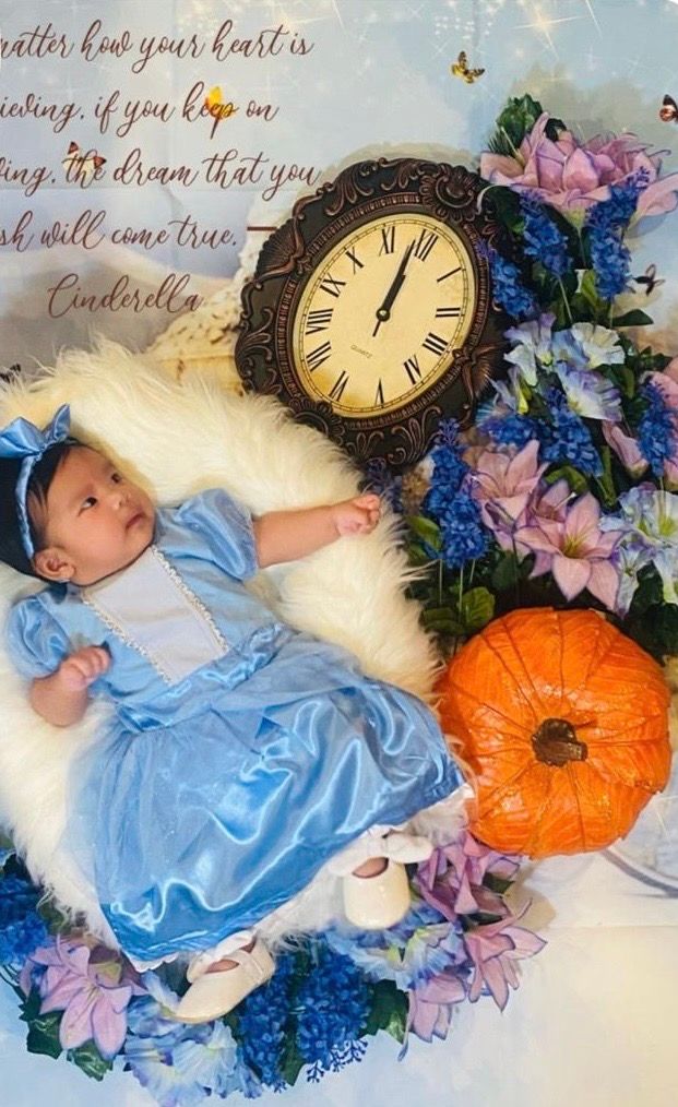 Baby dressed as Cinderella in blue gown surrounded by flowers, pumpkin, and clock in a fairy tale themed newborn photo.