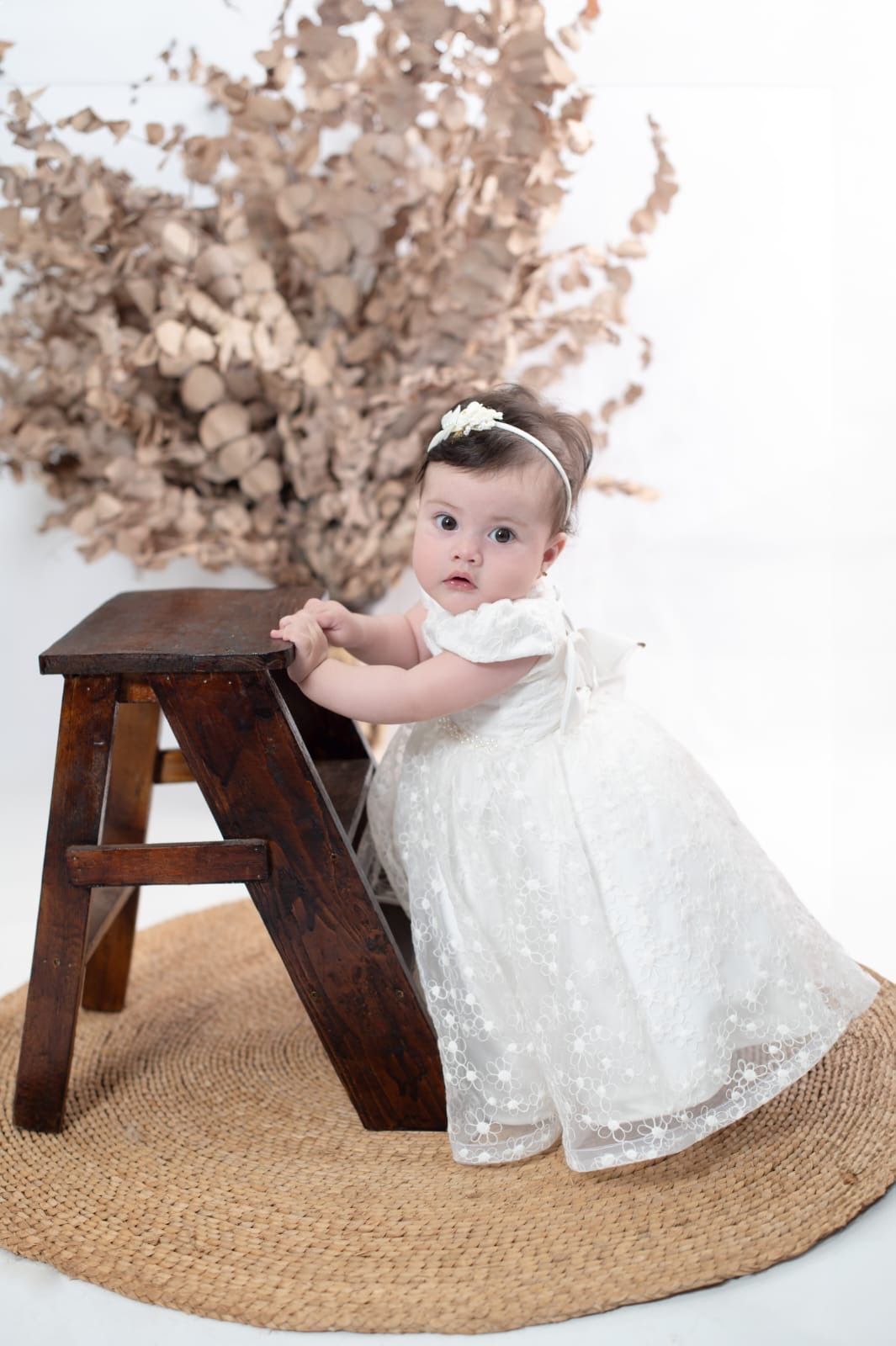 Baby girl in white lace dress stands by wooden step stool on woven rug with dried floral arrangement.