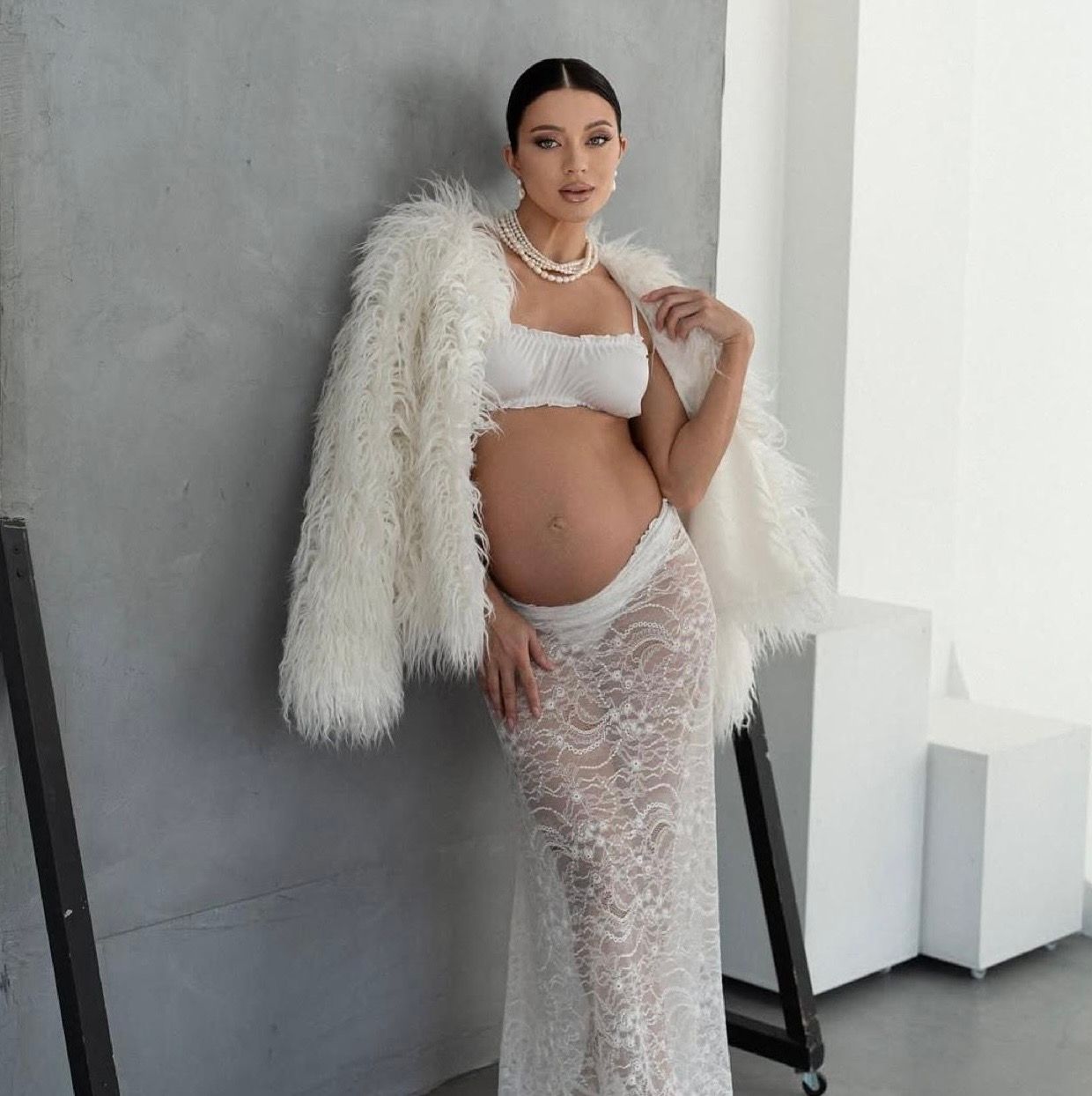 Pregnant woman posing in white lace skirt, crop top, pearl necklace, and fluffy fur coat against gray wall.