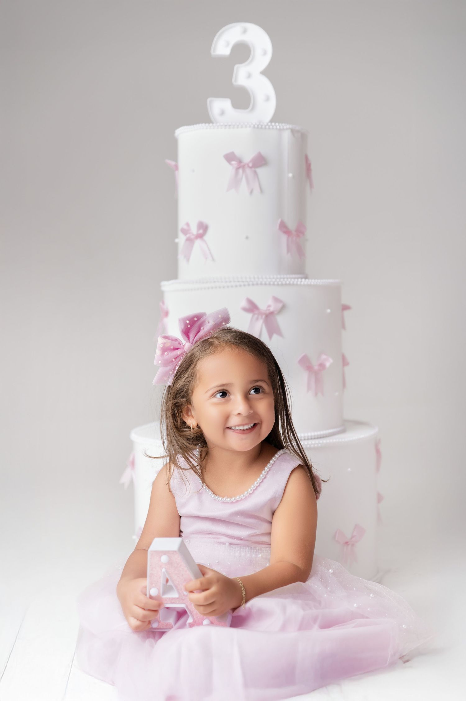 A smiling girl in a pink dress sits in front of a white 3-tier birthday cake decorated with pink bows.