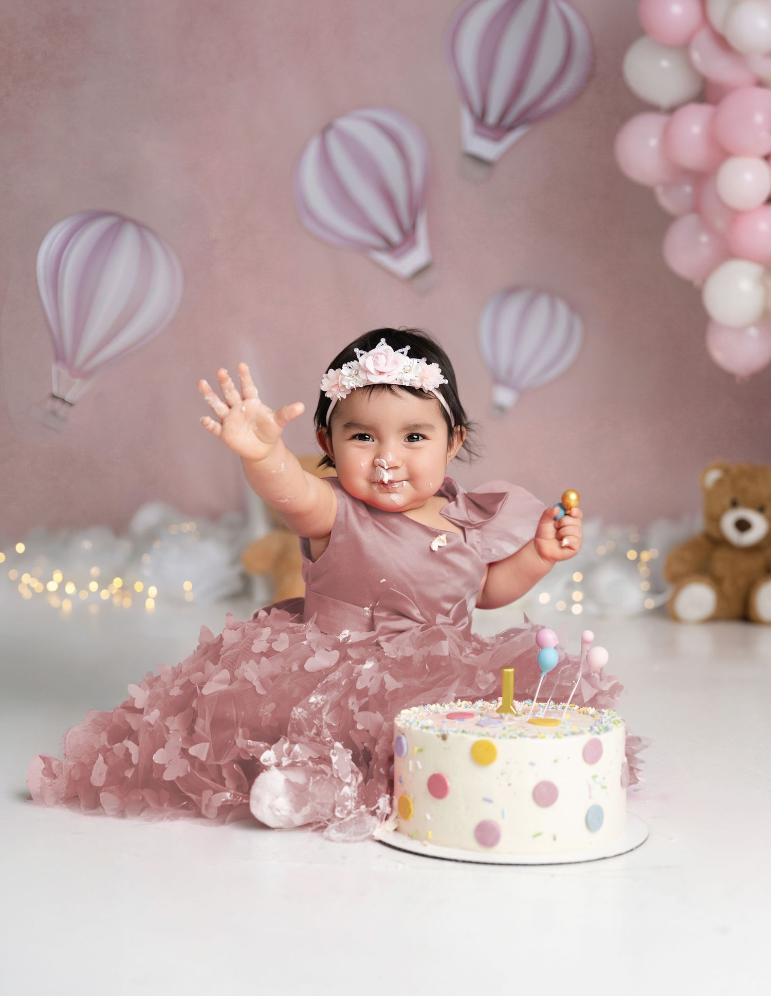 Baby girl in pink dress celebrates first birthday smashing a polka dot cake, surrounded by balloons and hot air balloon decor.