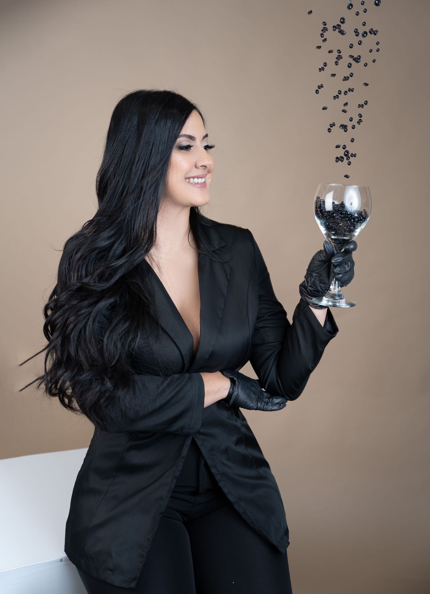 Professional photo of woman in black blazer holding a wine glass with sparkles floating above it against beige background.