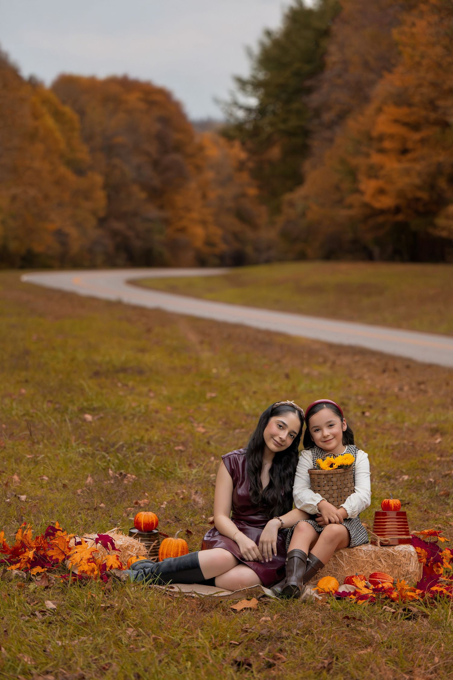 A mother and daughter sit together on hay bales surrounded by autumn fall decorations, pumpkins, and colorful foliage.