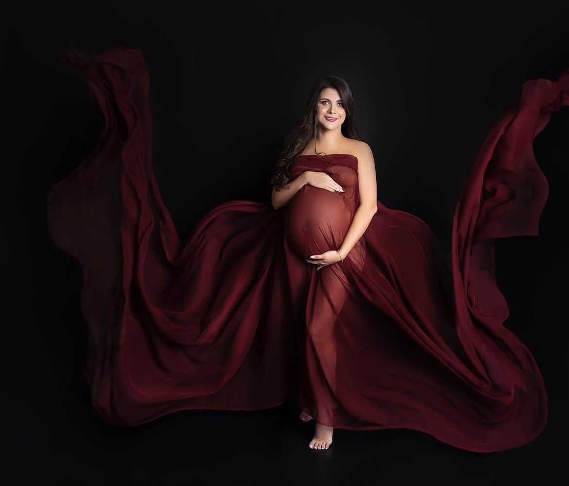 Pregnant woman in flowing red maternity gown with dramatic fabric movement against black studio background.