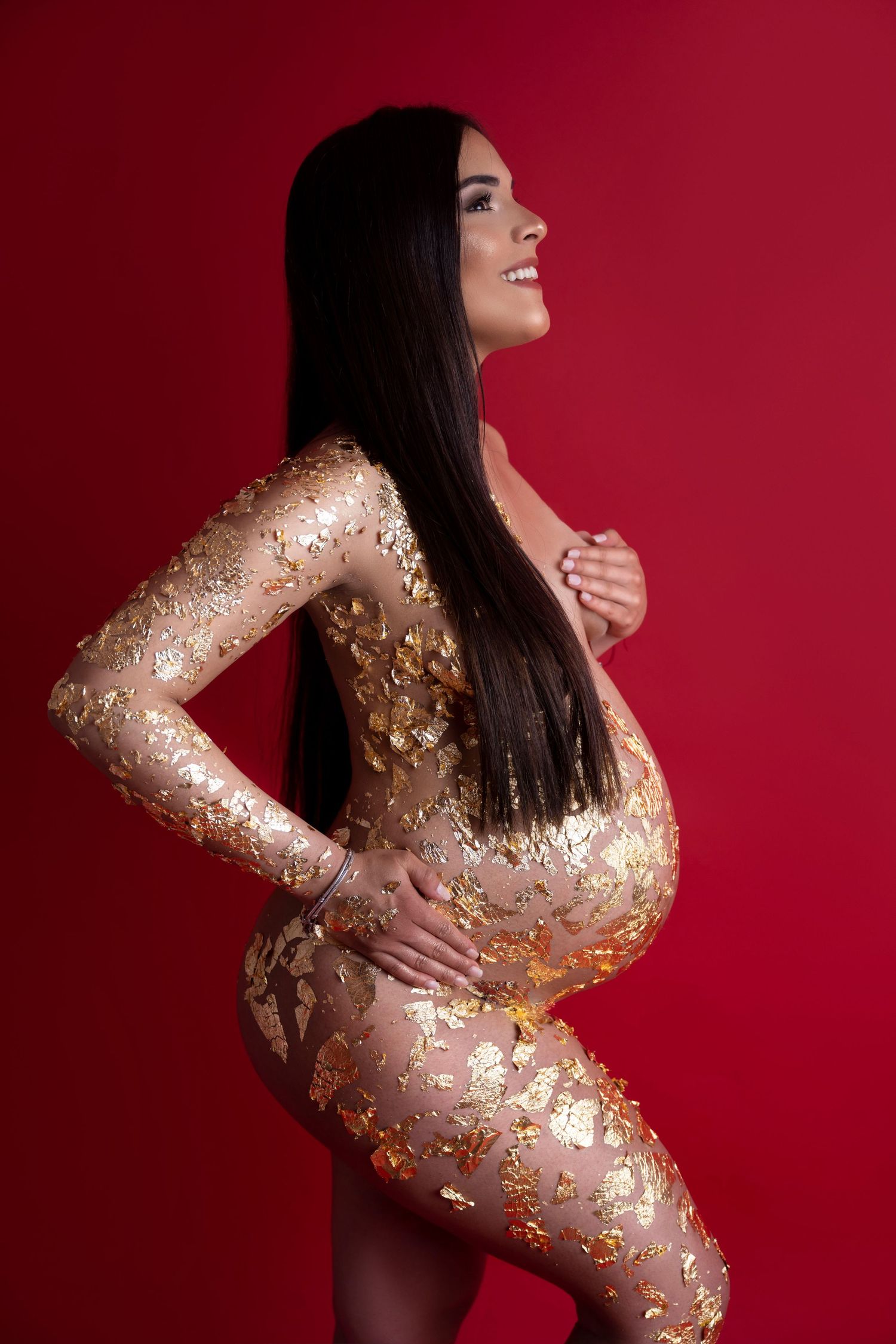 Artistic maternity portrait with gold glitter body art against red background.