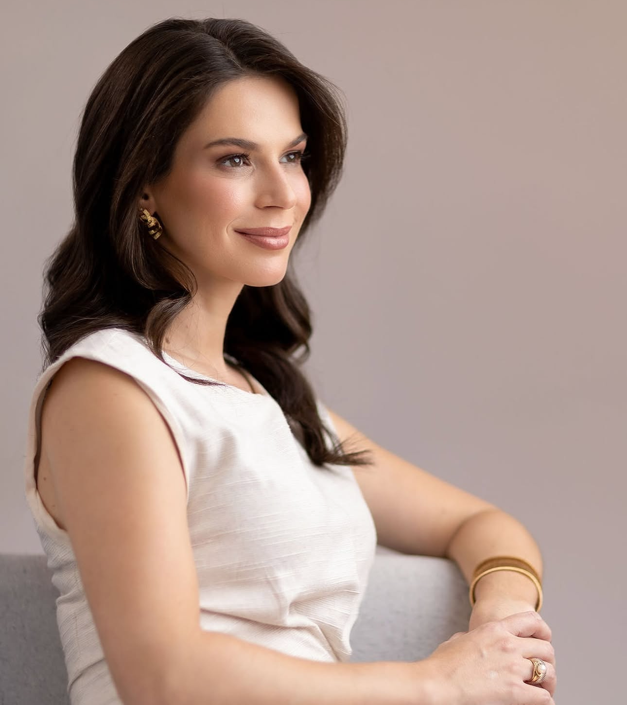 A confident woman with dark hair wearing a white sleeveless top and gold jewelry, seated and smiling gracefully.