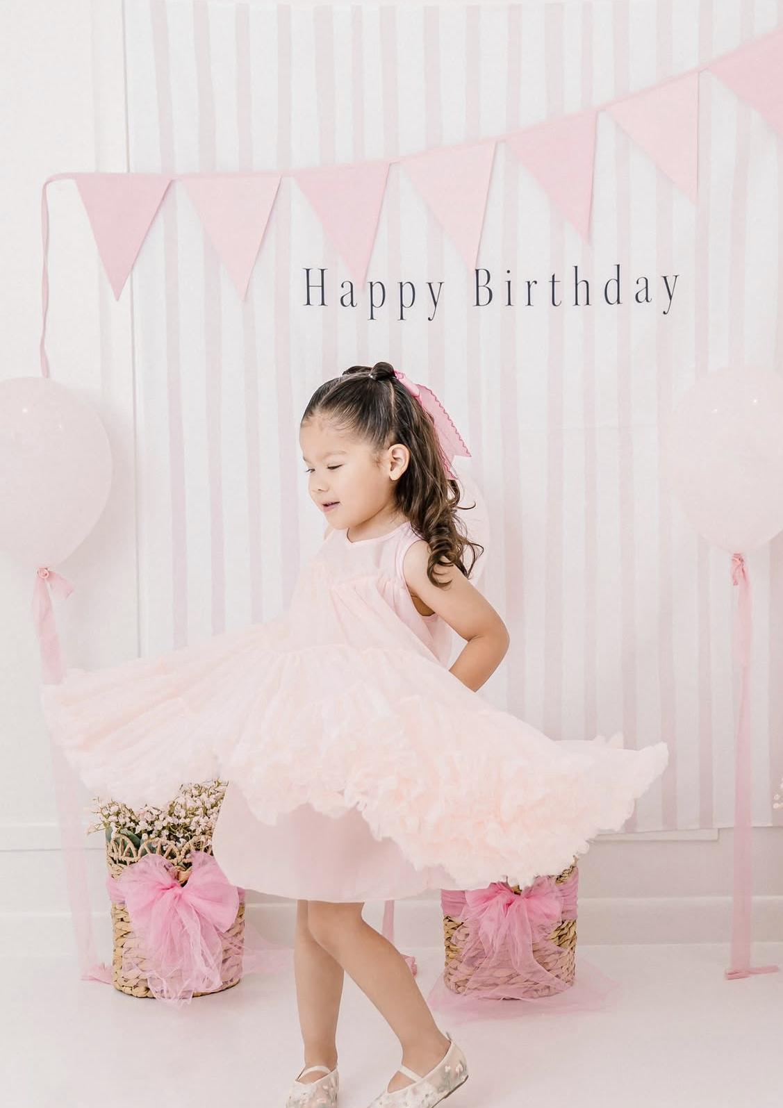 Young girl in pink tulle dress twirling at birthday party with pink pennant banner and decorative boxes.