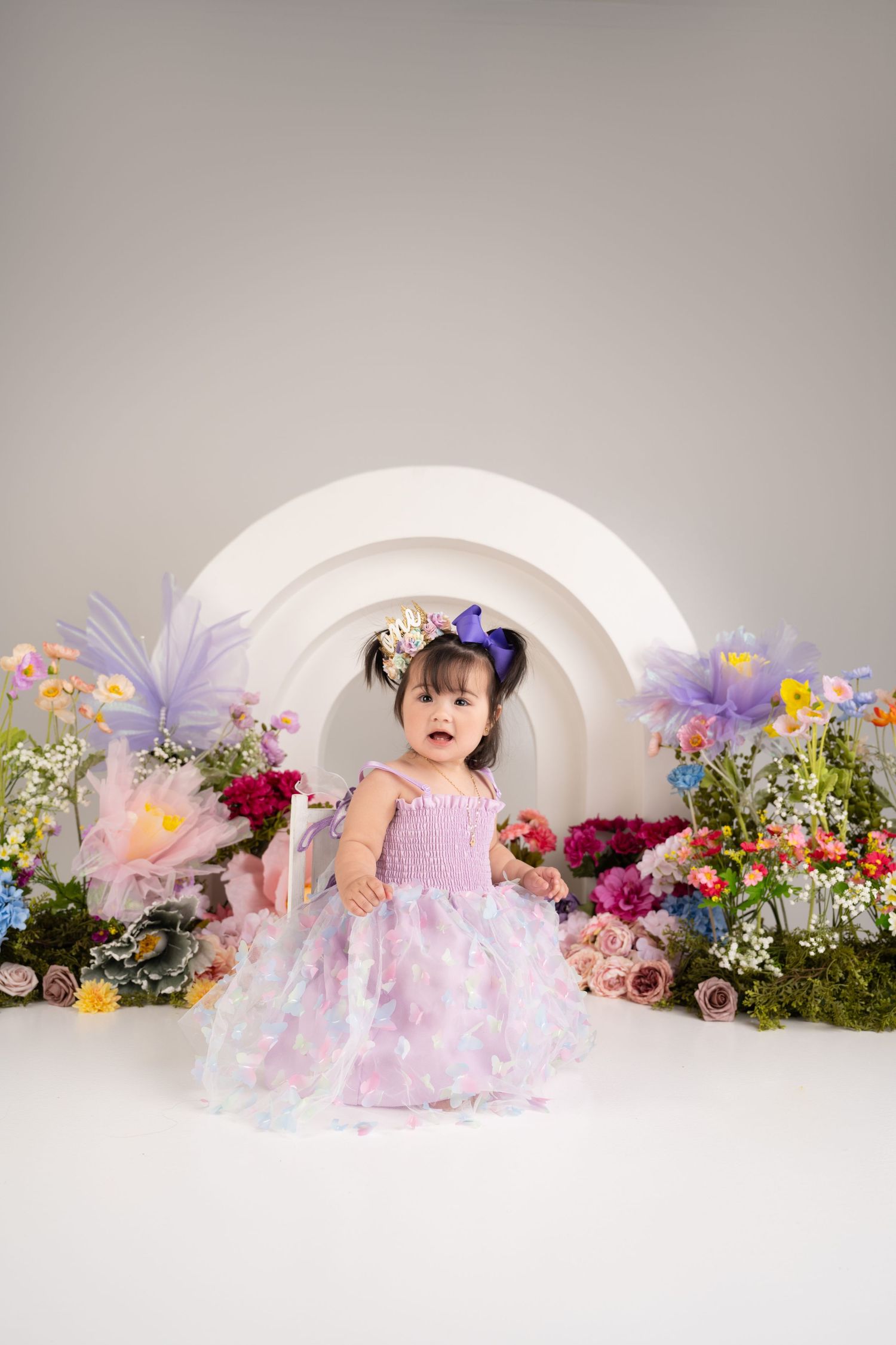 Child in princess dress stands in white arch decorated with colorful flowers.