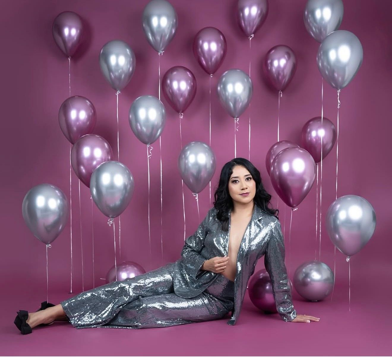 Woman in silver sequin suit poses on floor surrounded by pink and silver metallic balloons against pink backdrop.