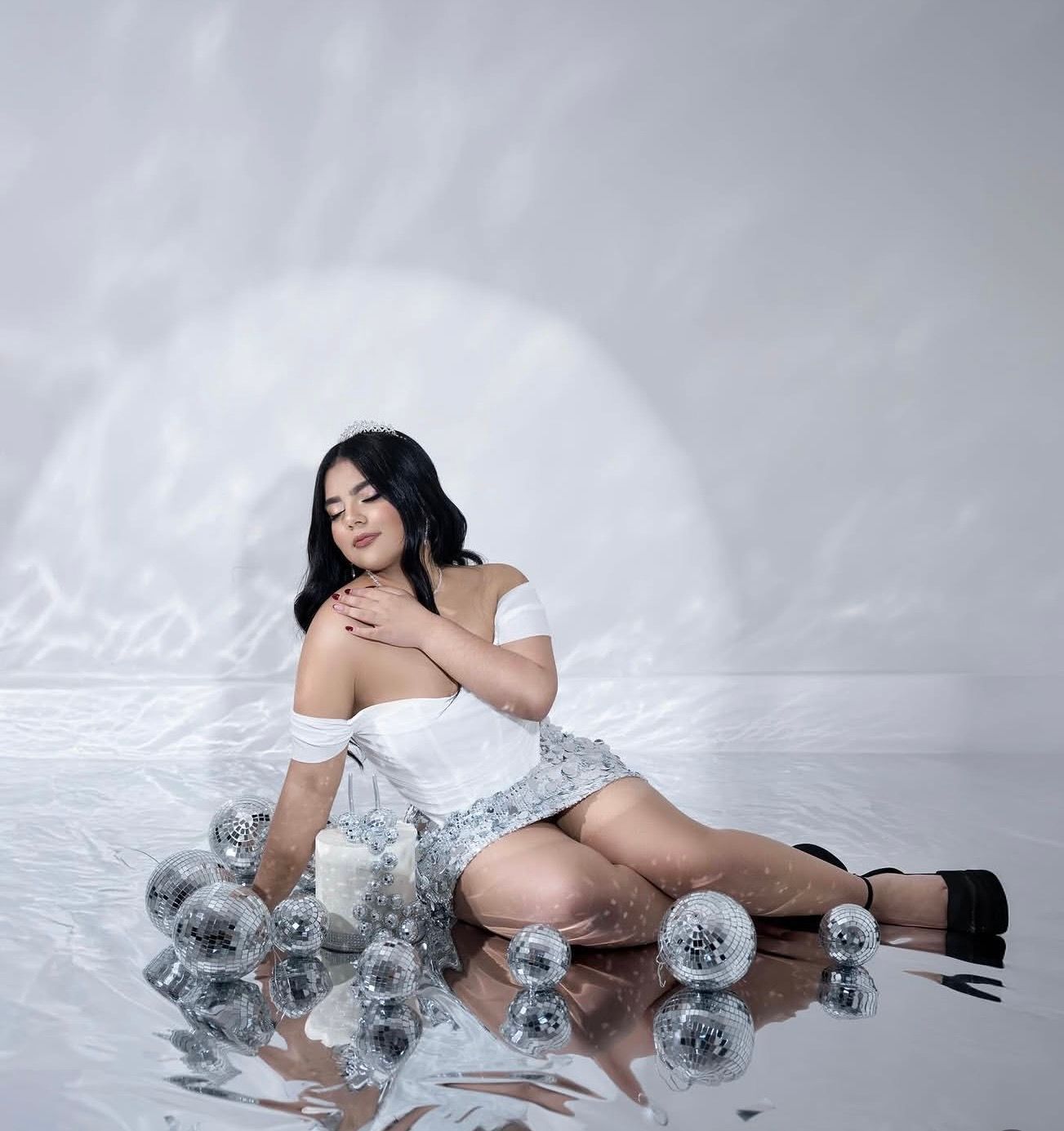 Woman in white off-shoulder top poses on reflective floor surrounded by silver disco balls in studio.