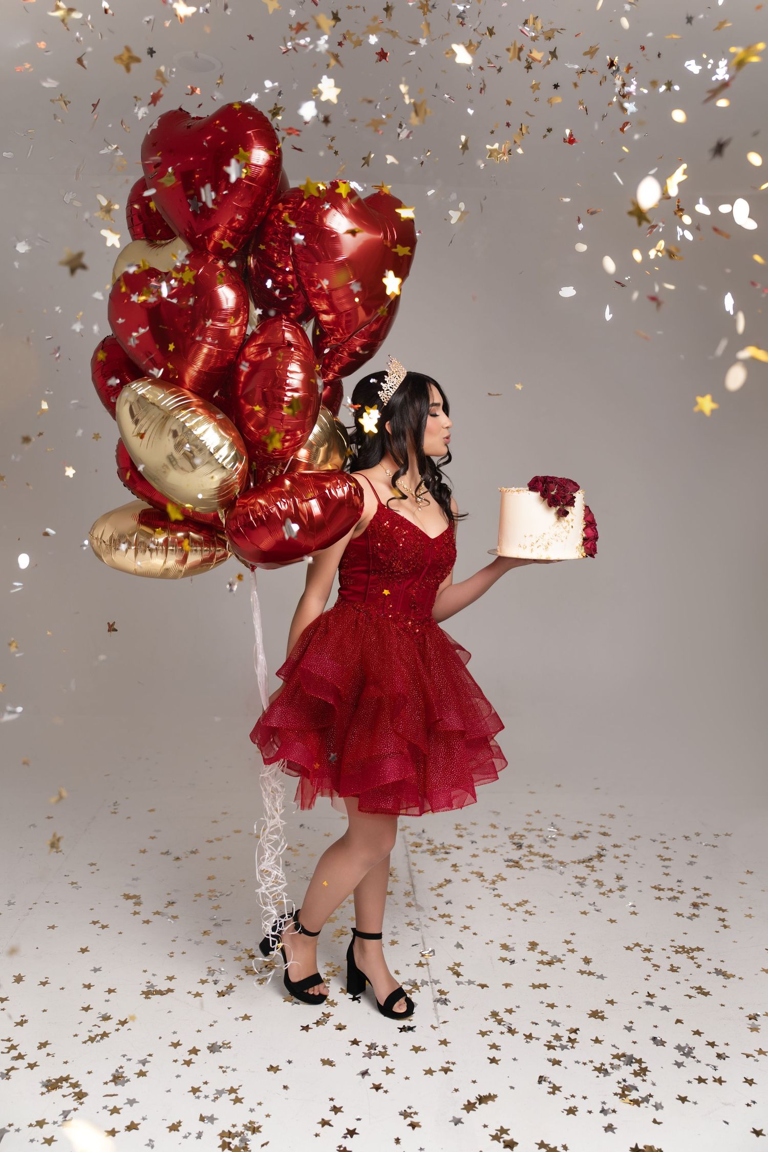 Woman in red dress holding heart balloons and cake with falling gold confetti at a birthday celebration.