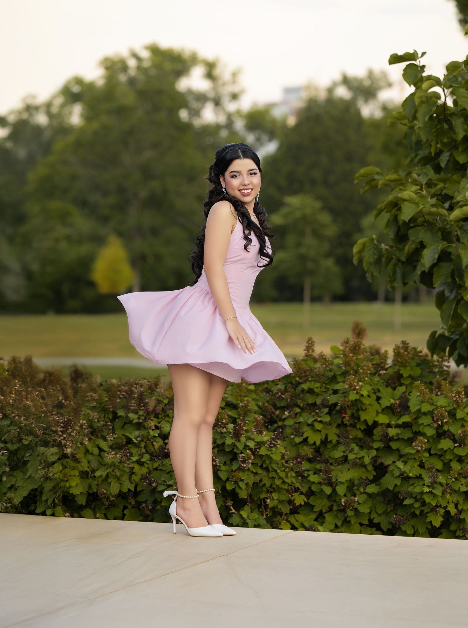 Young woman in pink dress posing outdoors, dress flowing in breeze, white heels, lush green park background.