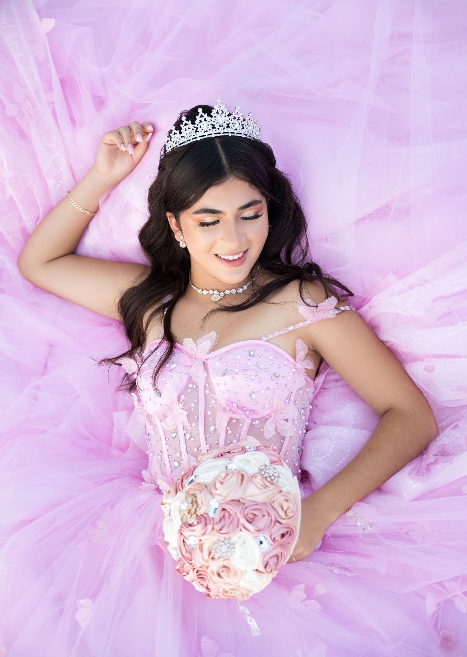 Young woman in pink quinceañera gown with tiara and floral bouquet lying on tulle skirt, smiling softly.
