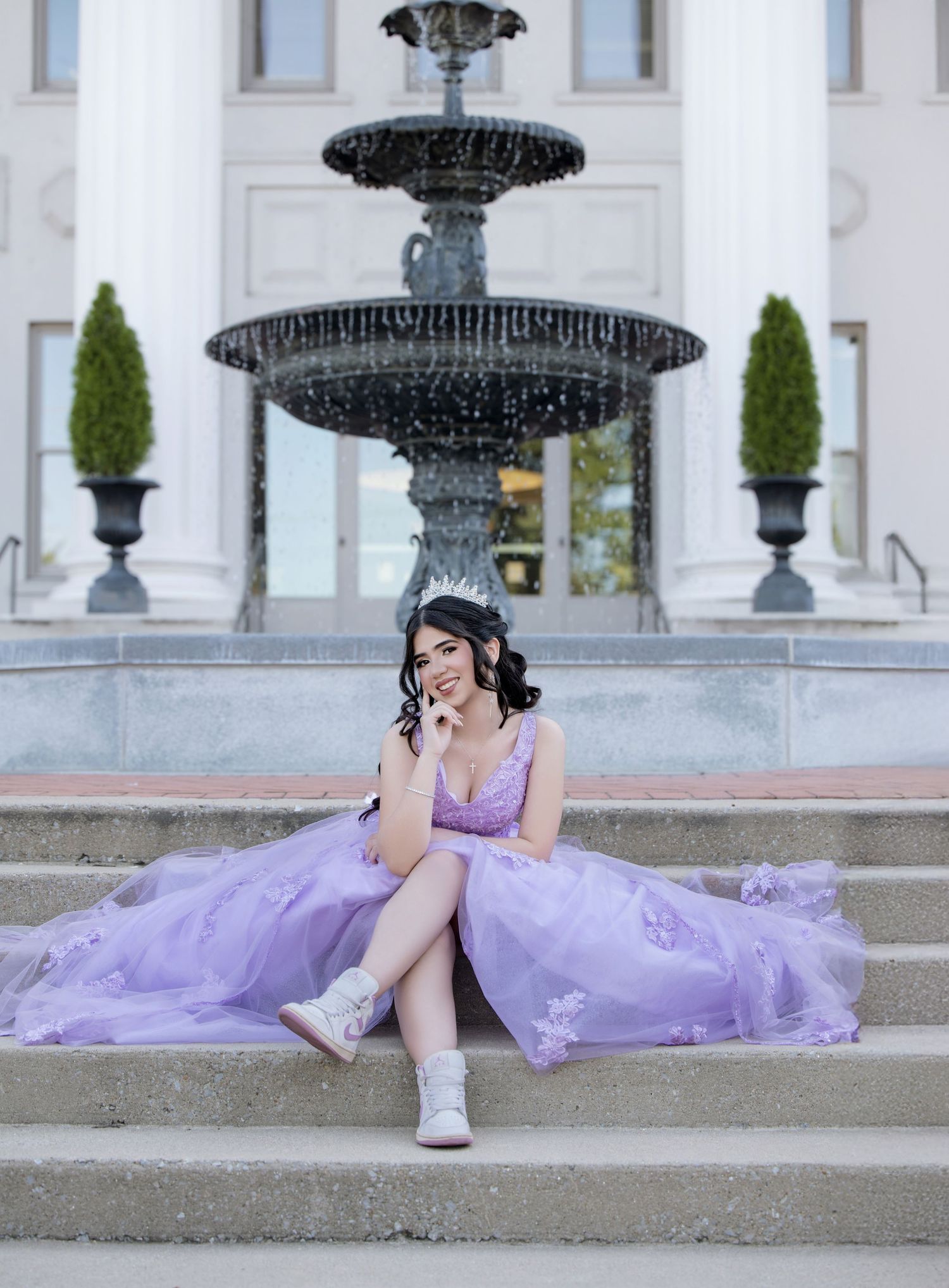 Young woman in lavender ball gown with tiara sits on fountain steps in elegant outdoor portrait.